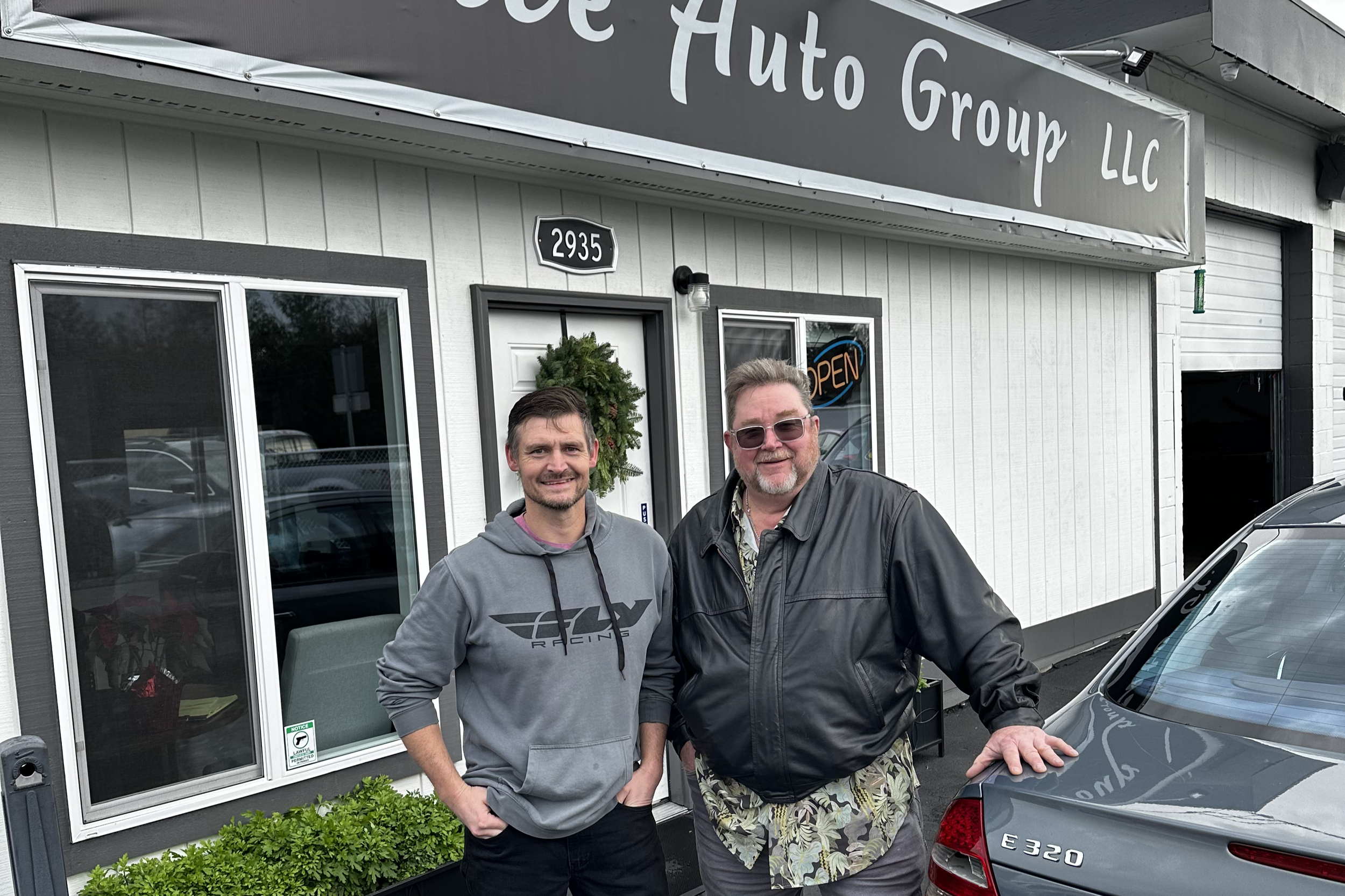 Two men standing outside of an auto group LLC dealership, smiling, with a building behind them that has a sign and large windows. One man is wearing a gray hoodie, and the other man is in a black jacket and sunglasses. A silver Mercedes-Benz E 320 is parked nearby.