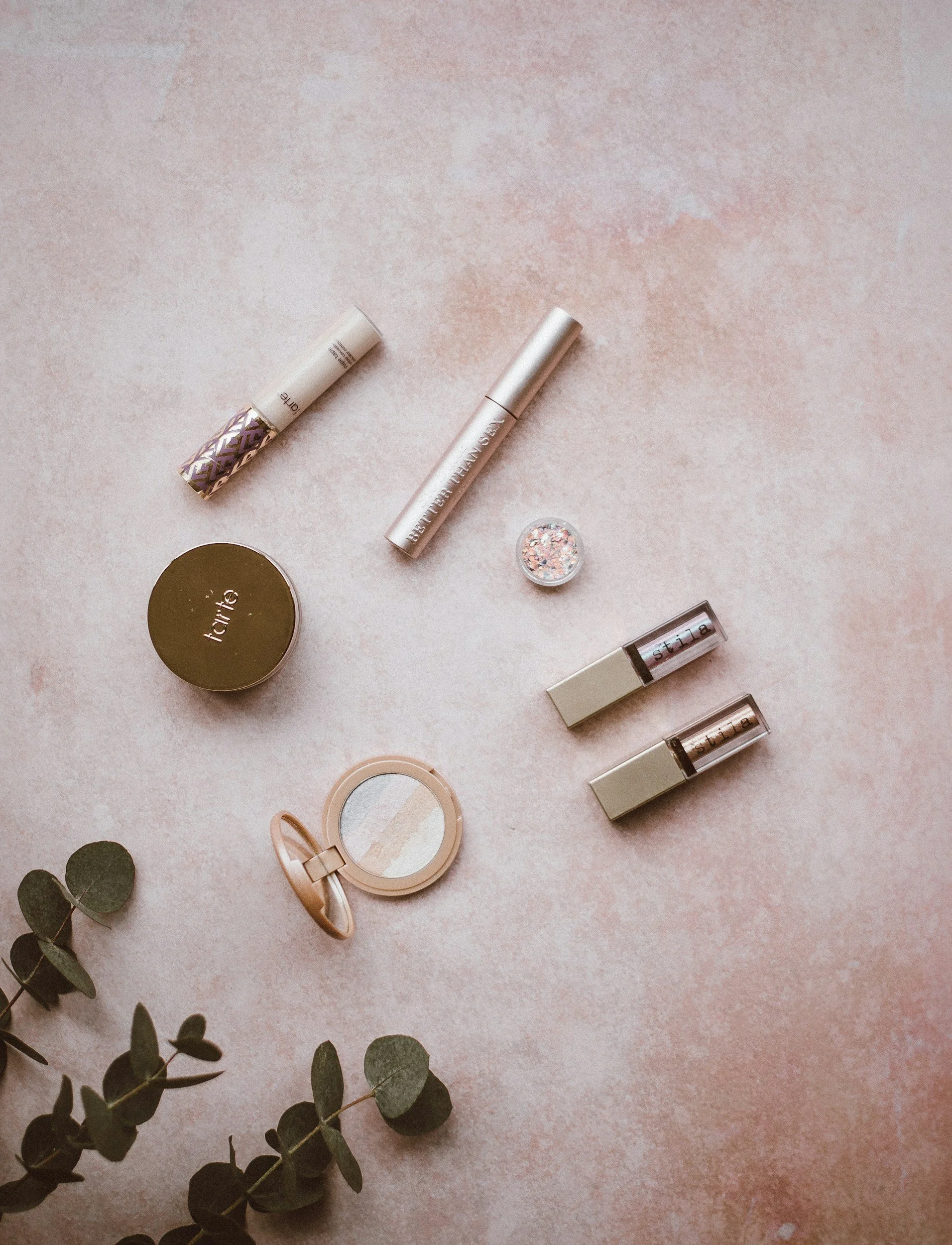 Beauty products, including lip glosses, a small round mirror, a compact with blush or powder, a container of glitter, and a sprig of eucalyptus on a pink textured background.