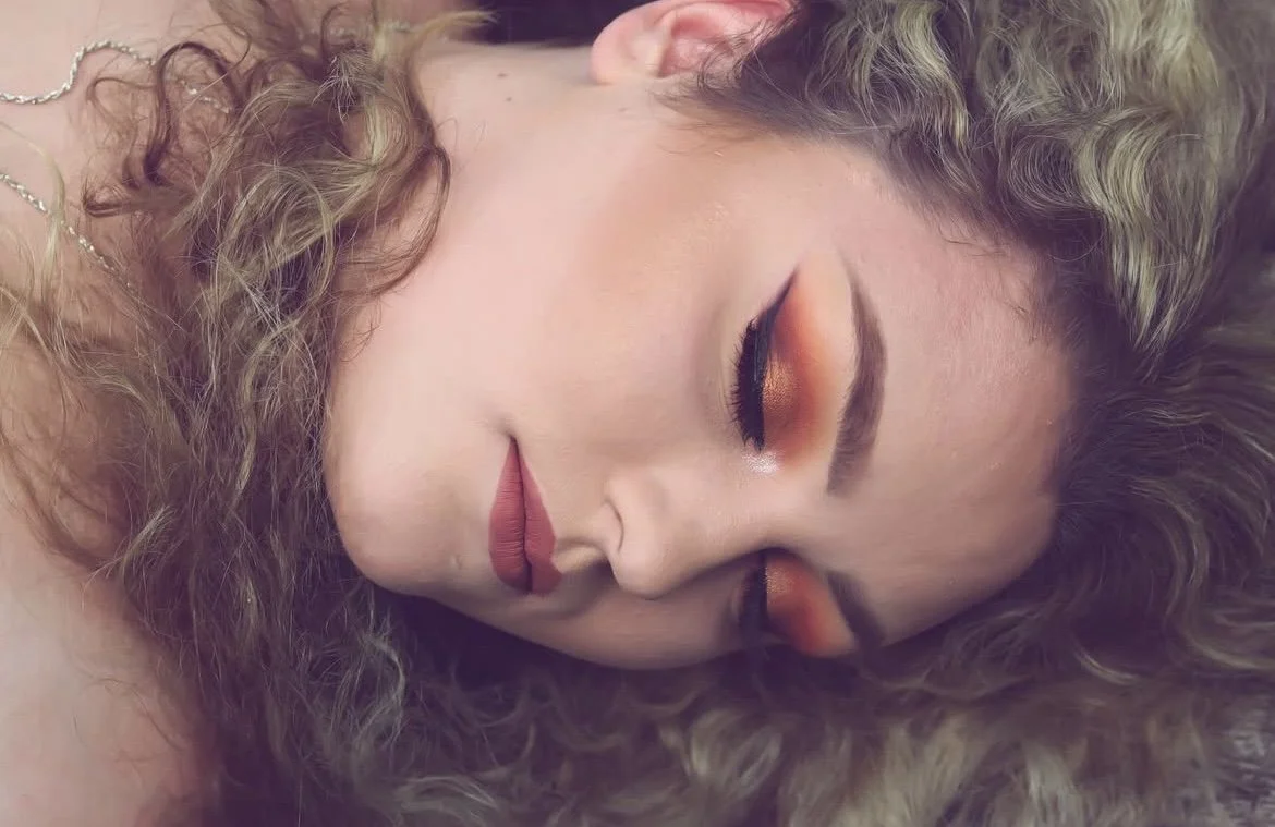 A woman with curly hair and makeup with orange and gold eyeshadow, resting with eyes closed.