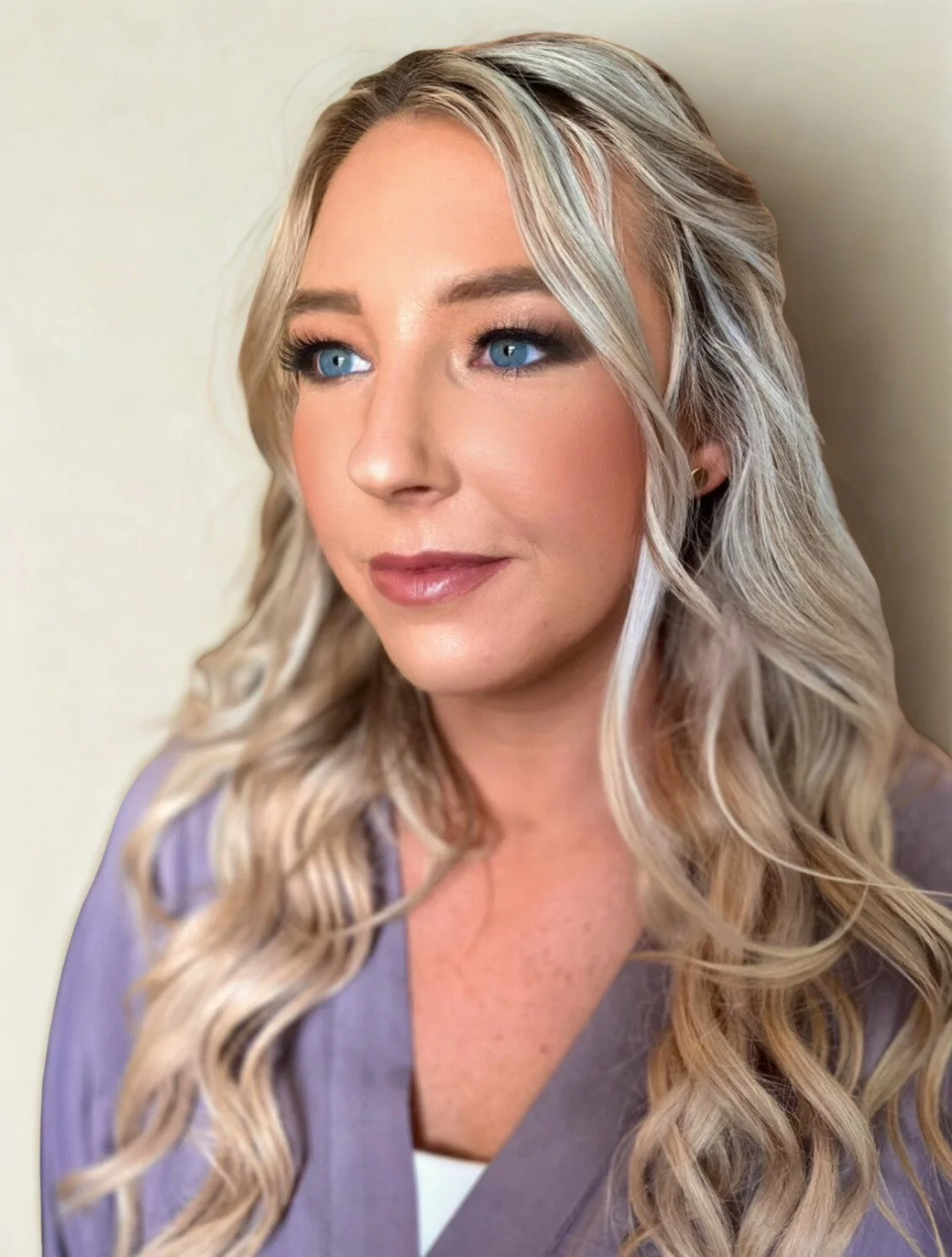 Close-up portrait of a woman with long blonde wavy hair and blue eyes, wearing a lavender blazer, against a plain background.
