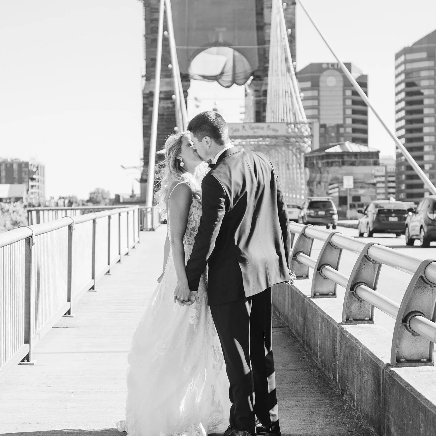 A black-and-white photo of a bride and groom sharing a kiss on a city bridge during daytime, with city buildings and parked cars in the background.