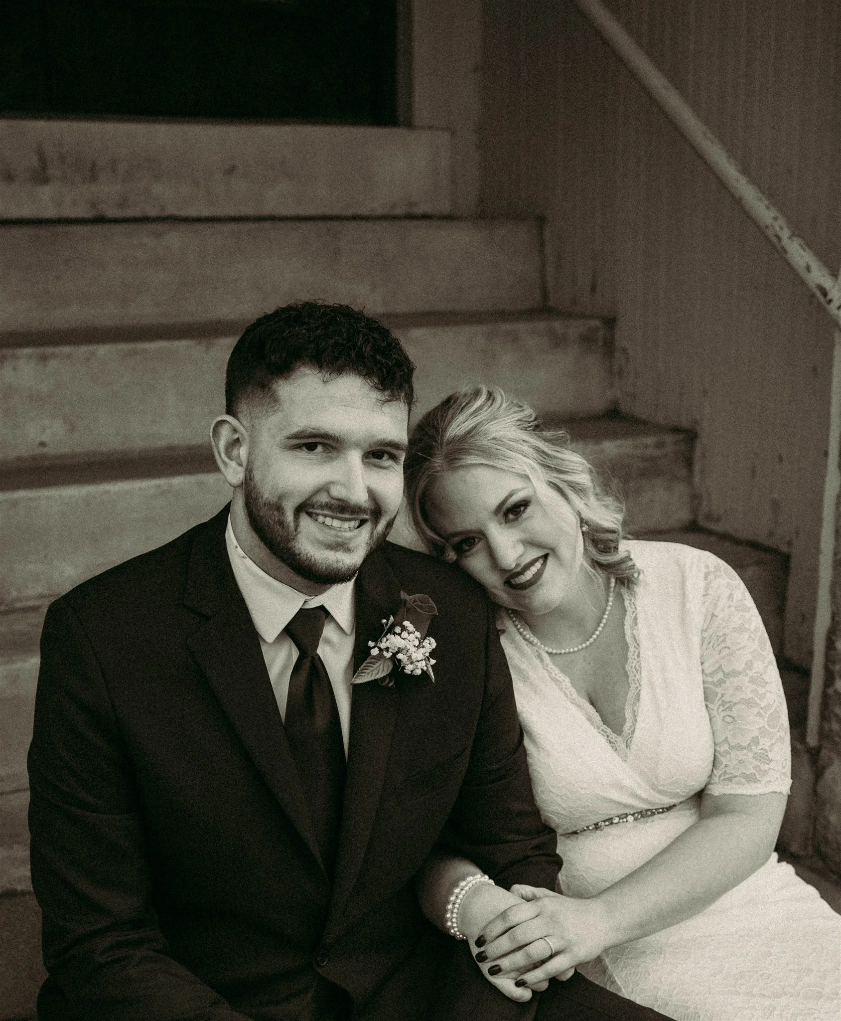 A black and white photo of a man and woman sitting close together, smiling. The man is wearing a suit and tie with a flower boutonniere, and the woman is wearing a lace dress and pearl necklace.