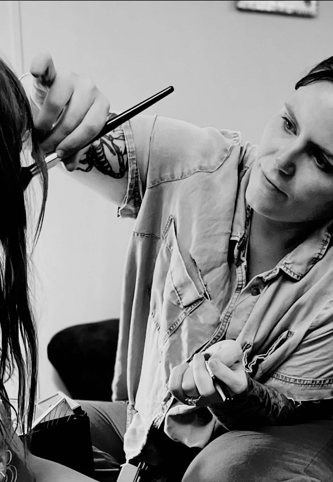 A man with tattoos on his arm is tattooing a client's arm. The client has long hair, and the man is focused on his work in a tattoo studio.
