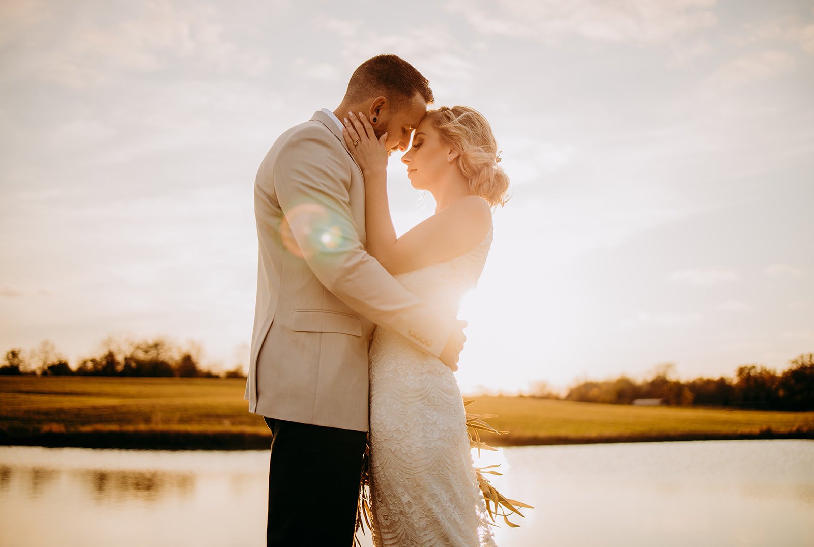 A newlywed couple embracing each other at sunset by a lake, with the sun shining behind them.