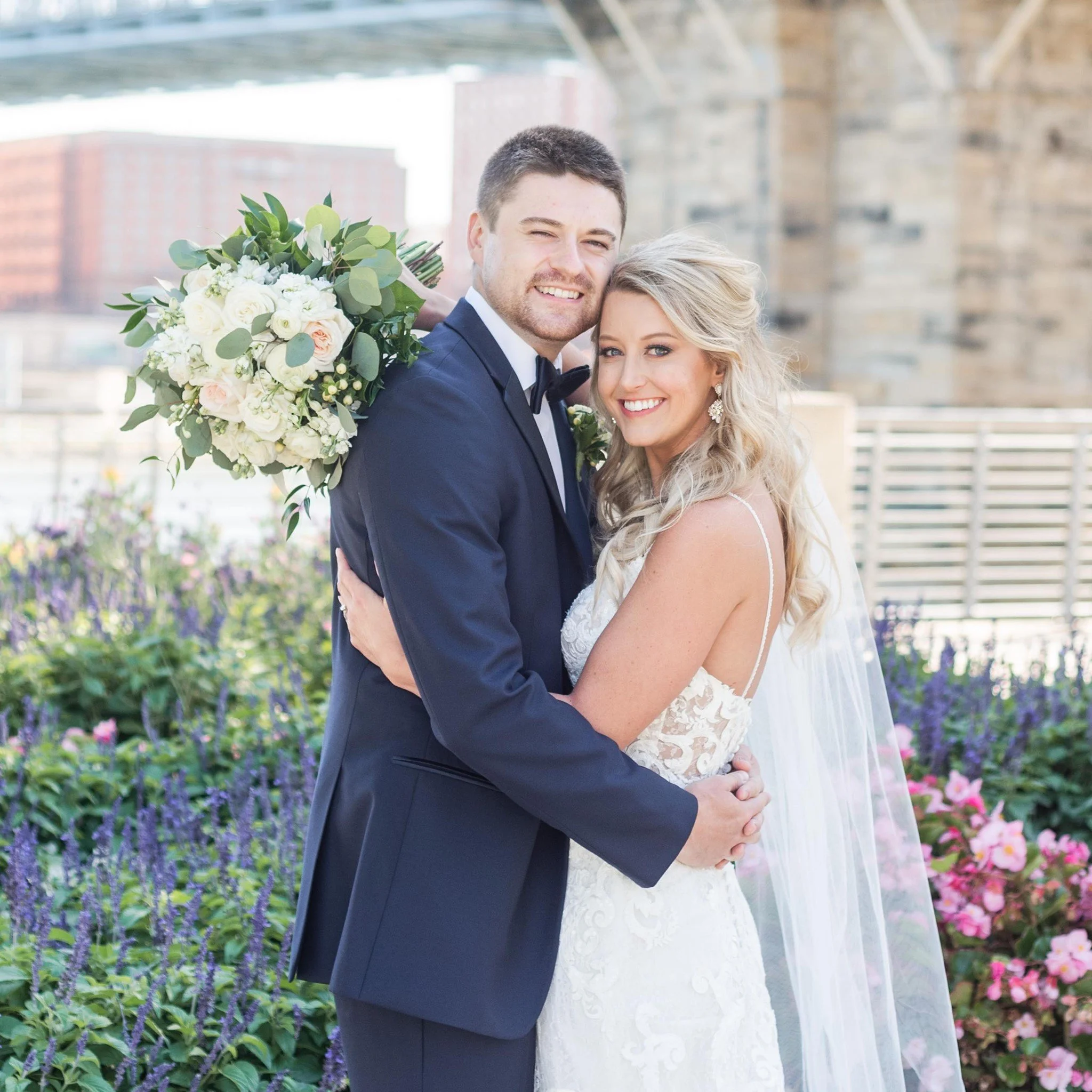 A happy bride and groom embracing on a rooftop garden with flowers, the groom holding a bouquet of white and pink flowers.