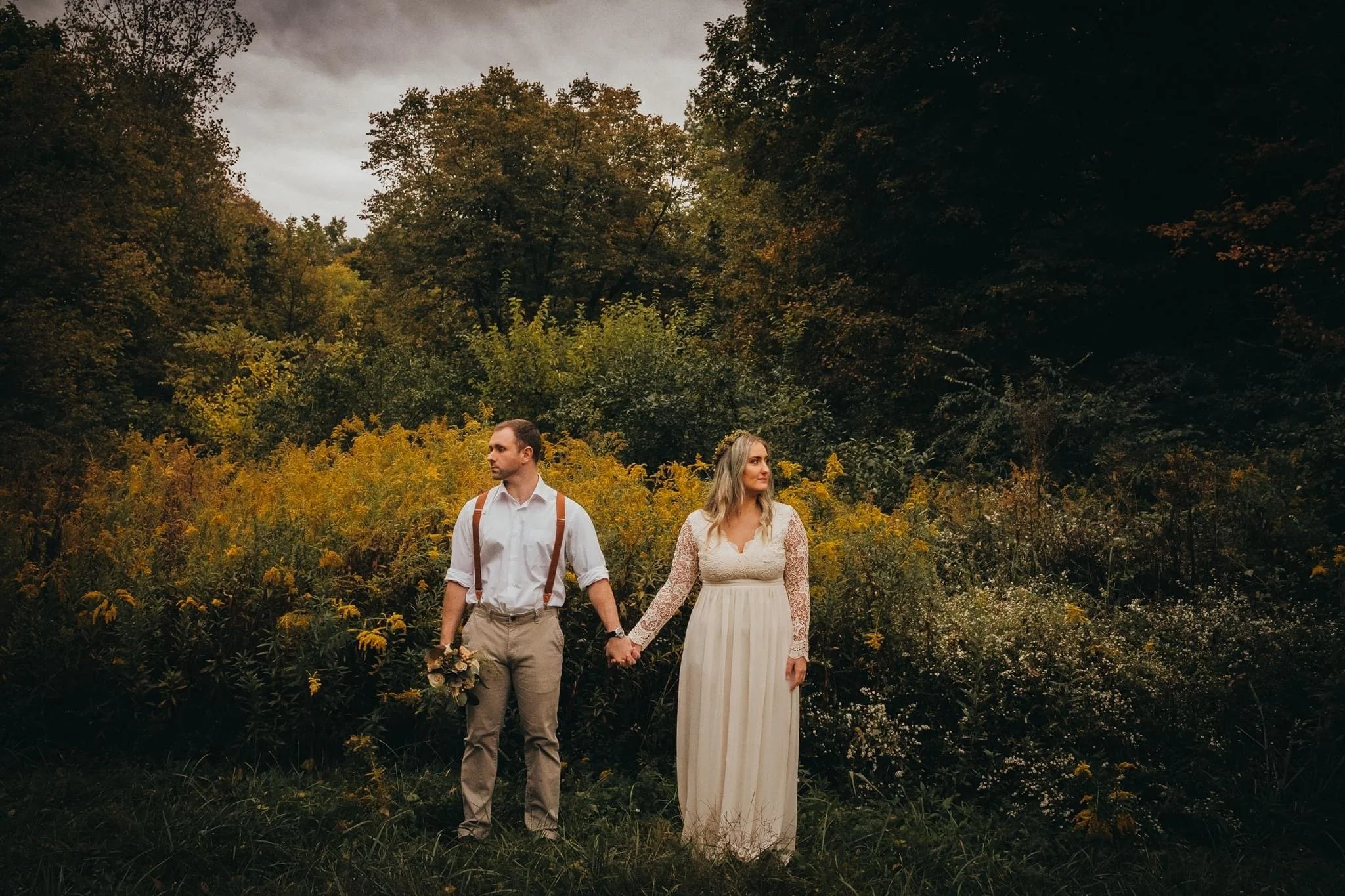 A couple holding hands in a forest during the daytime. The woman is wearing a long white dress with lace sleeves and the man is dressed in beige pants, a white shirt, and suspenders. The man holds a bouquet of flowers.