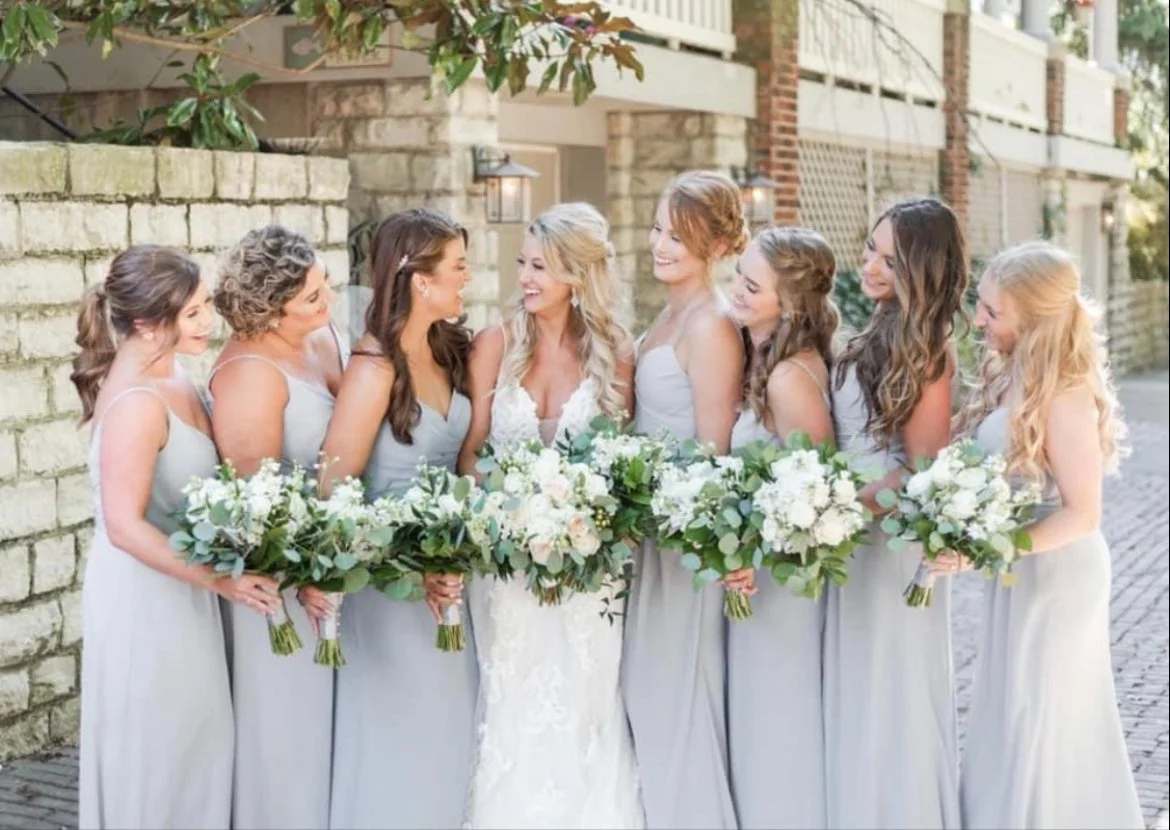 Bride and seven bridesmaids standing outdoors, smiling and holding bouquets of white and green flowers, in front of a brick wall.