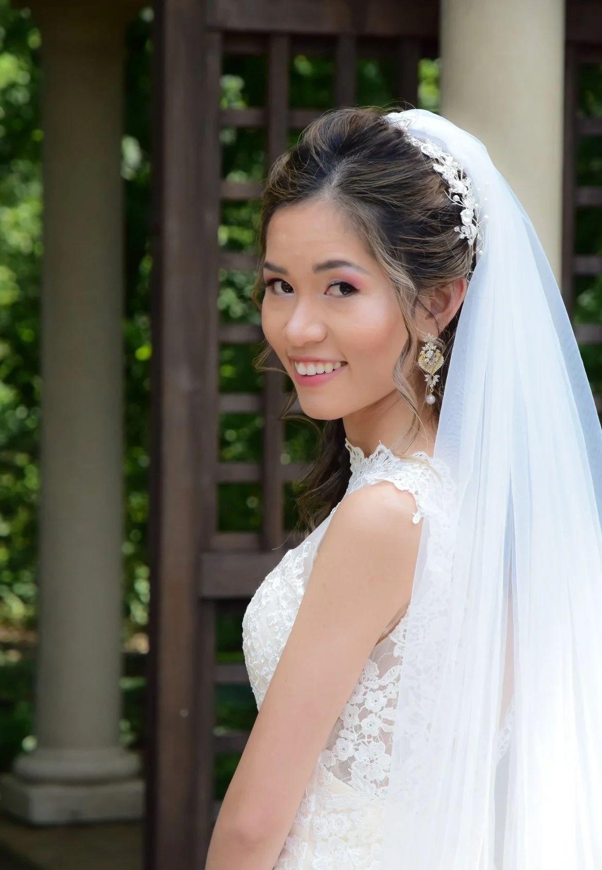 A smiling bride in a lace wedding dress with a veil and floral hair accessory, standing outdoors next to a wooden structure and green foliage.