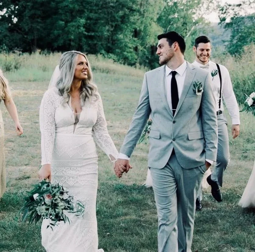 A bride and groom holding hands at an outdoor wedding ceremony, with the bride in a white lace dress holding a bouquet and the groom in a light gray suit, surrounded by greenery and smiling guests.