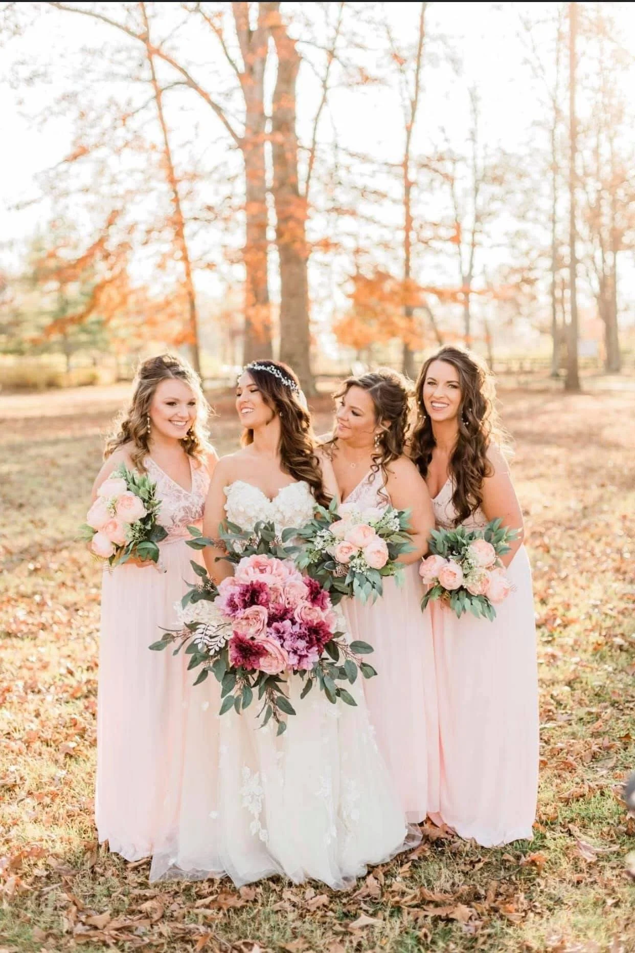A bride in a white wedding dress with her four bridesmaids dressed in pink standing outdoors during autumn, holding bouquets of pink and white flowers with green foliage, surrounded by fall foliage and trees.
