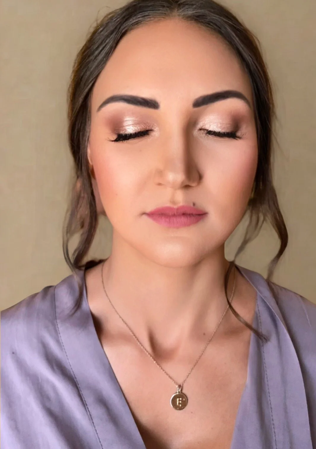 A woman with closed eyes, wearing makeup, a satin purple top, and a gold necklace with an 'E' pendant.