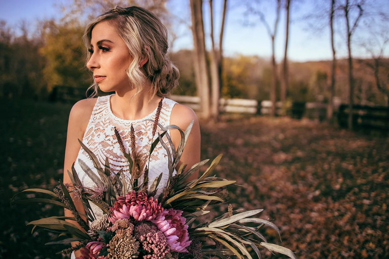 A woman with blonde wavy hair, wearing a white lace dress, holding a bouquet of pink and purple flowers and greenery, outdoors with trees and a wooden fence in the background during autumn.