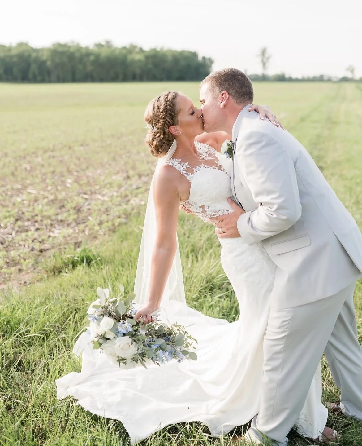 A bride and groom share a kiss outdoors in a grassy field with a farm background, the bride holding a bouquet of white and blue flowers.