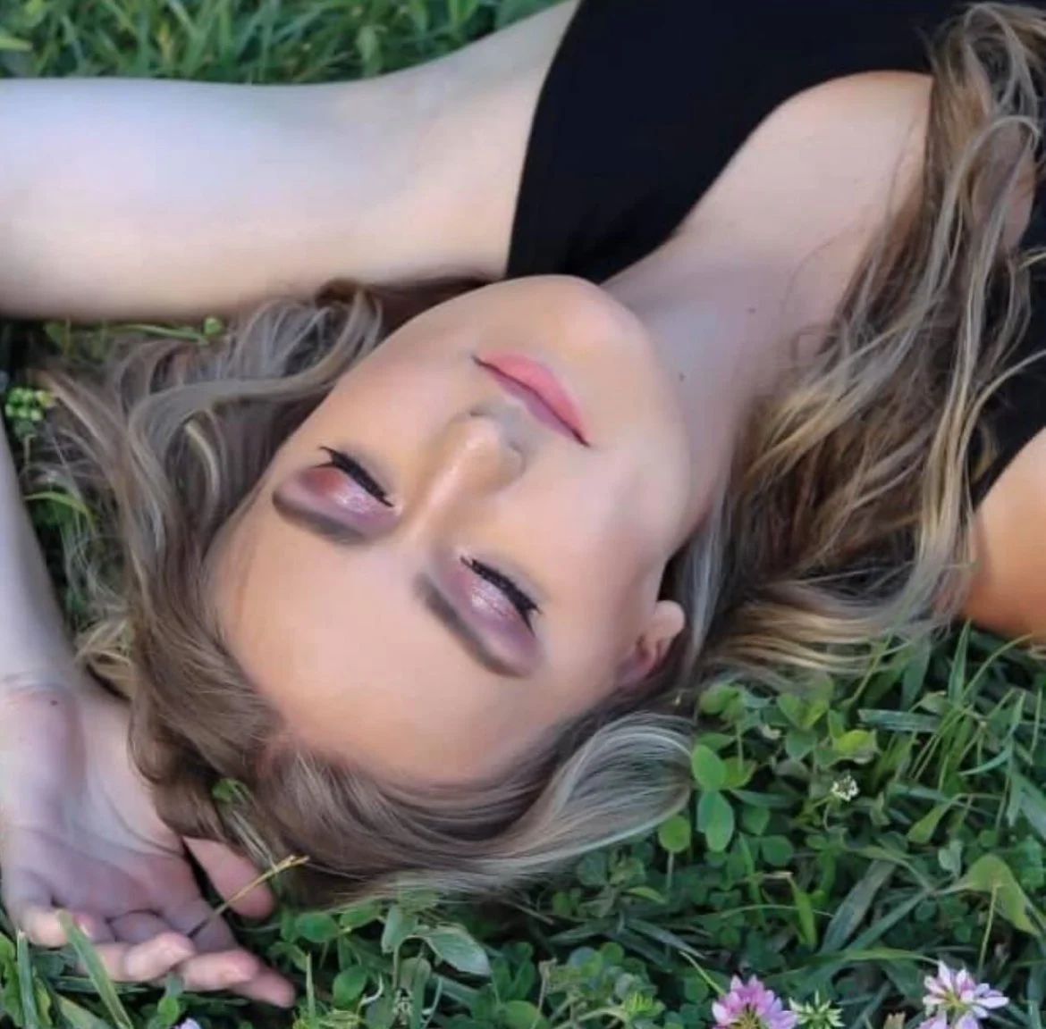 A woman lying on grass with her eyes closed, wearing a black top and having wavy blonde hair.