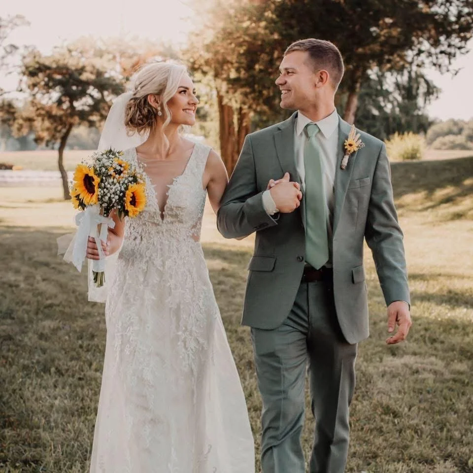 A bride and groom walking outdoors on a sunny day, holding hands and smiling at each other. The bride is wearing a lace wedding dress and holding a bouquet of sunflowers, while the groom is dressed in a gray suit with a light green tie and boutonniere.
