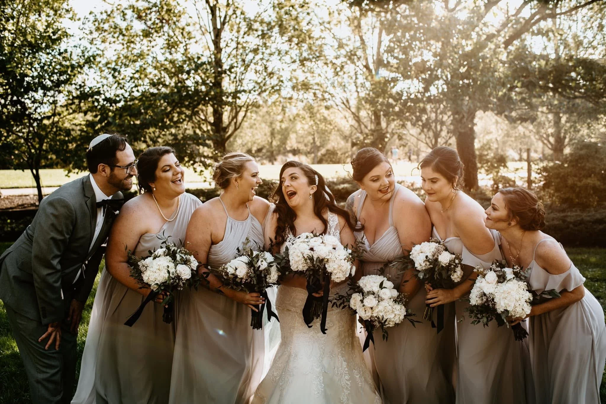 Group of women in bridesmaid dresses and one man in a suit, all smiling and laughing, holding bouquets, gathered outdoors in a park during sunset.
