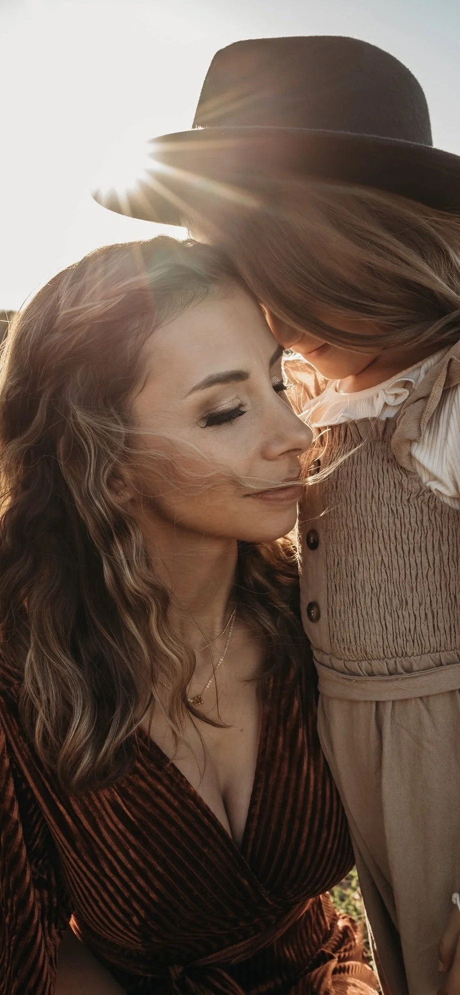 A woman with long, wavy hair and makeup, wearing a deep V neckline dress, and a child with long hair wearing a beige vest and white blouse, sharing an intimate moment outdoors during sunset.