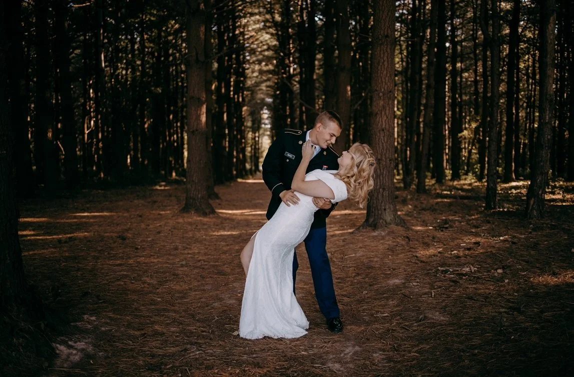 A couple in wedding attire dancing in a forest with tall trees and a natural ground surface.