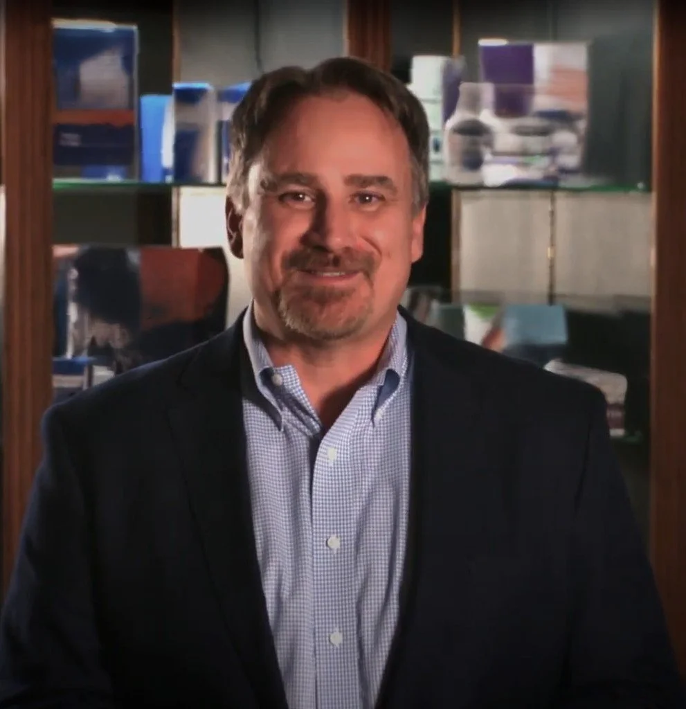 A middle-aged man with short brown hair, a beard, and light skin, wearing a dark blazer and a light blue, checkered dress shirt, standing in front of a glass display case filled with medication and medical supplies.