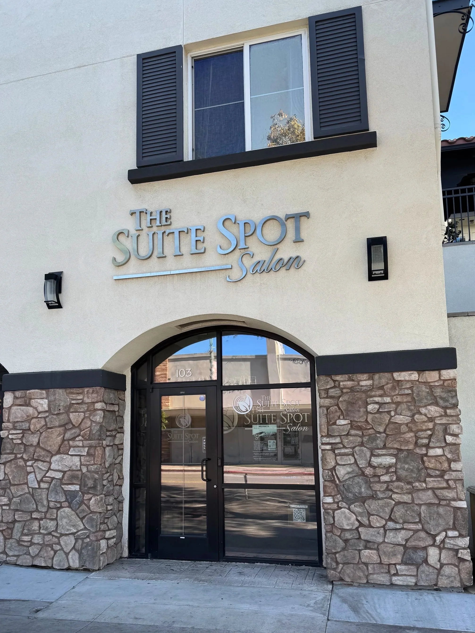 The exterior of a salon named 'The Suite Spot Salon' with a stone facade, glass door with logo, and a window above with black shutters.
