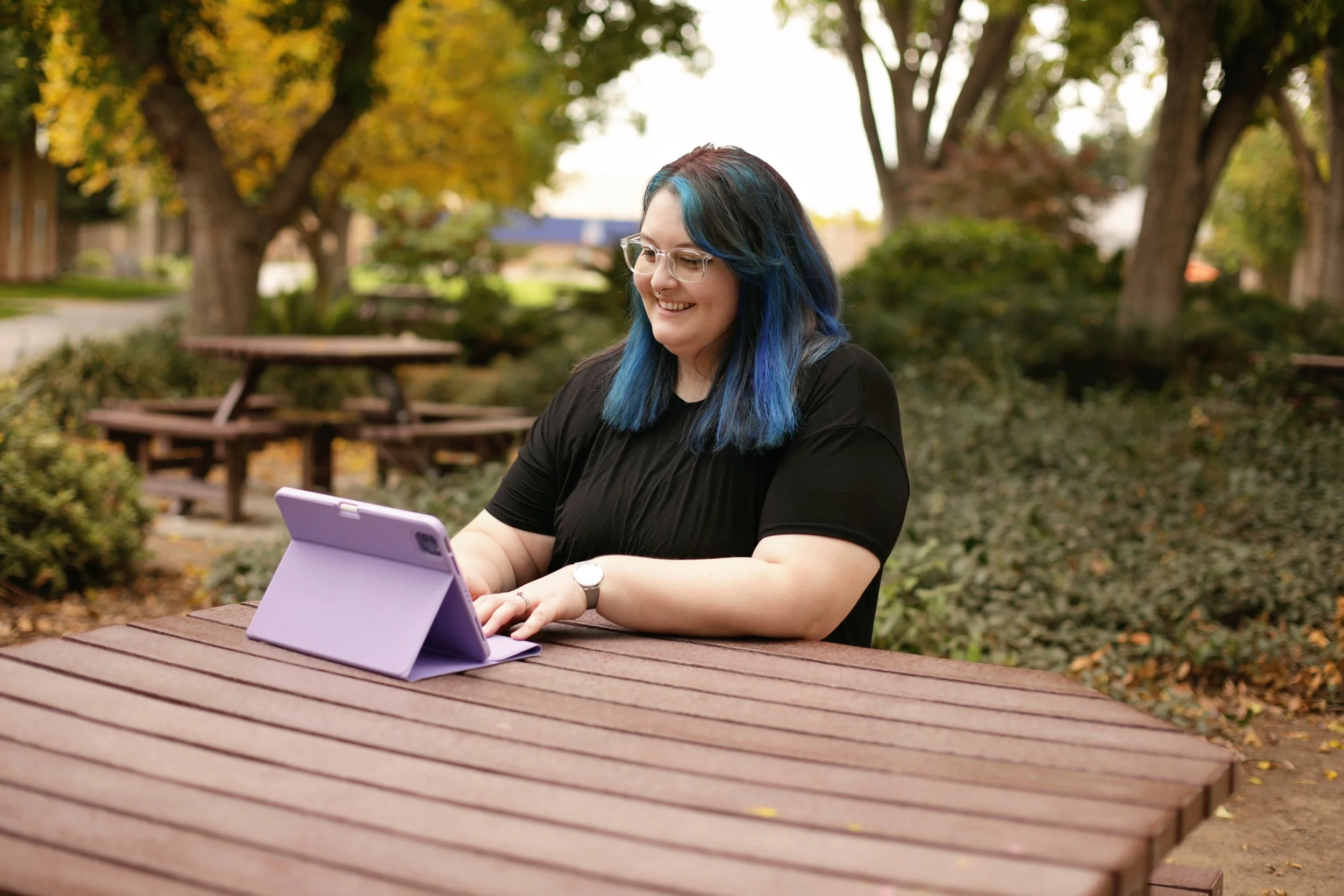 A woman with blue and black hair, wearing glasses and a black shirt, is sitting at a wooden picnic table outdoors in a park, using a purple tablet.