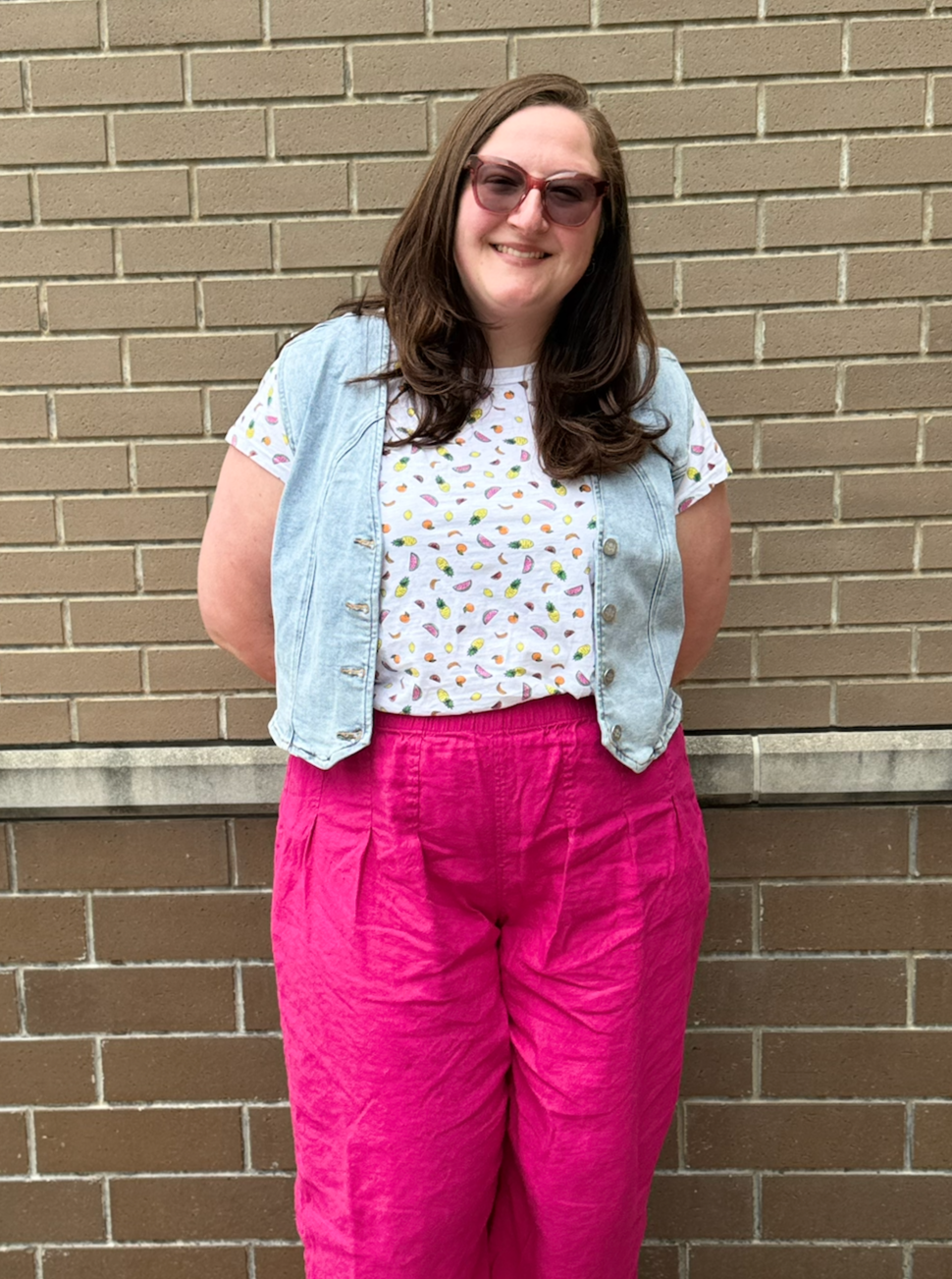 A woman with long brown hair wearing pink sunglasses, a white T-shirt with a colorful pattern, a light blue denim vest, and bright pink pants, standing in front of a beige brick wall, smiling.