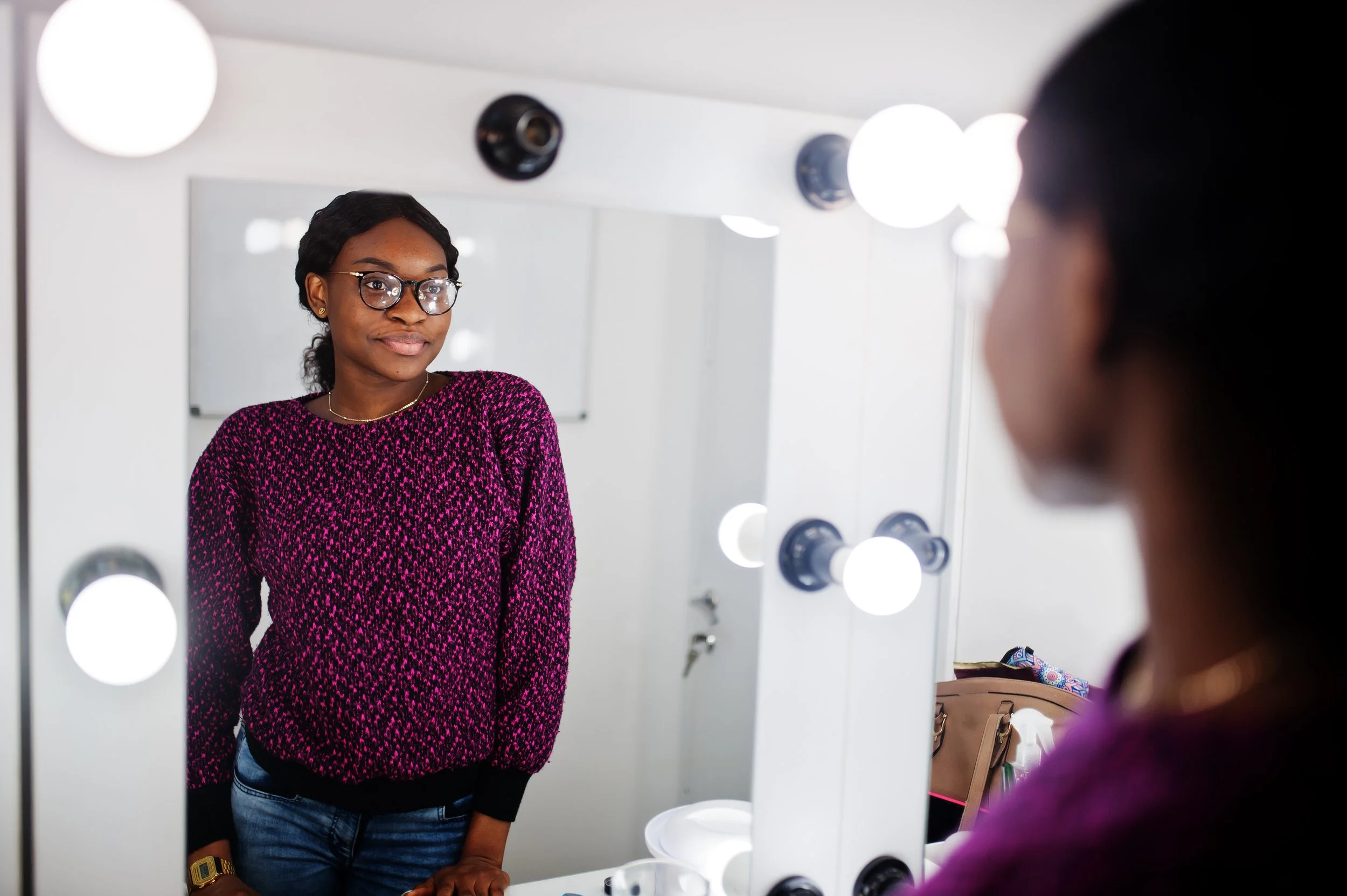 A woman in a pink and black speckled sweater, wearing glasses, looking at her reflection in a mirror with bright lights.