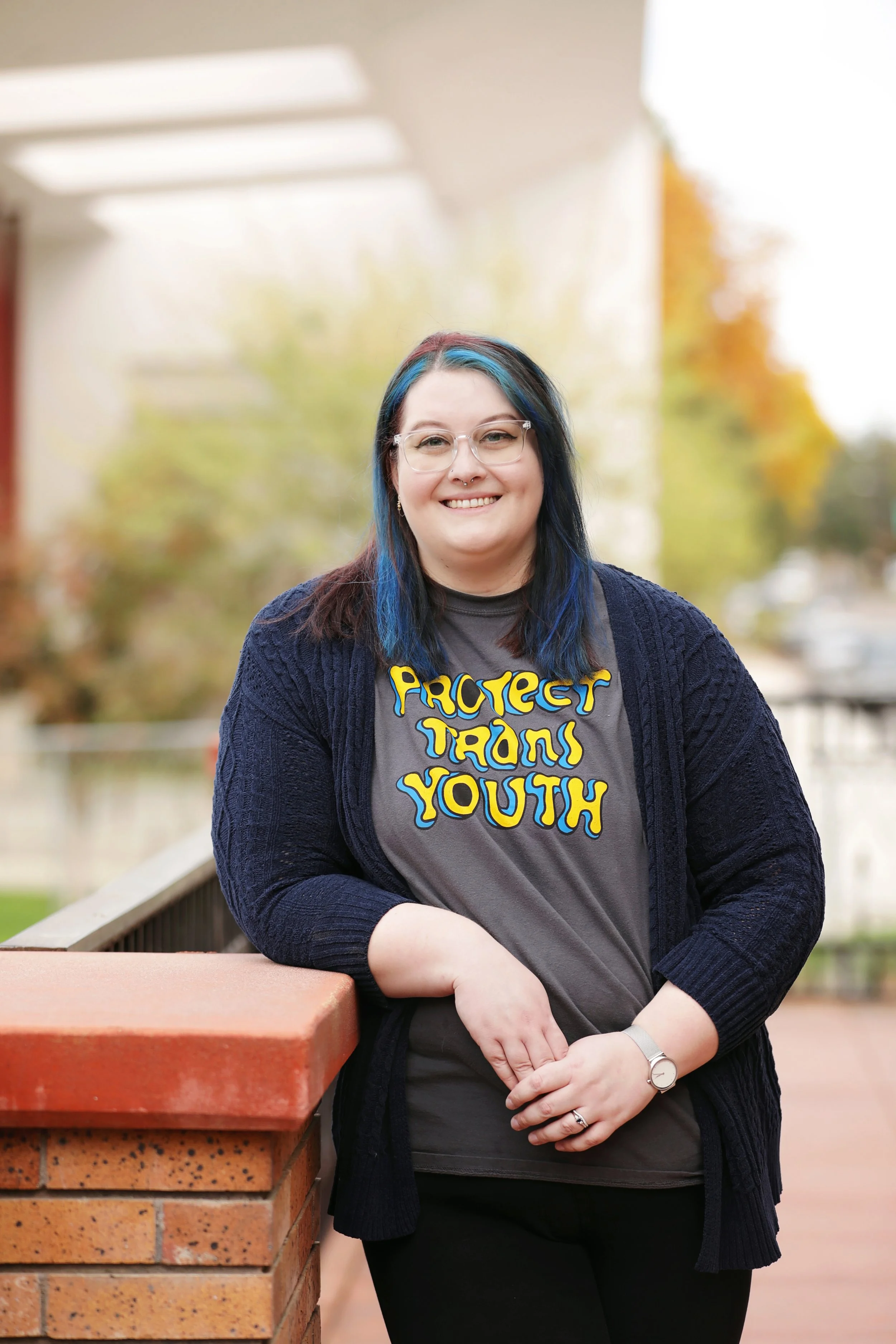 A woman with blue and black hair, glasses, and a septum piercing, smiling and leaning on a brick railing outside with autumn trees in the background. She is wearing a gray t-shirt with yellow and blue text that says 'PROTECT TRANS YOUTH' and a dark cardigan.