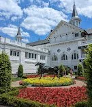 White church with steeple, surrounded by colorful flowers and trees, under a partly cloudy sky.