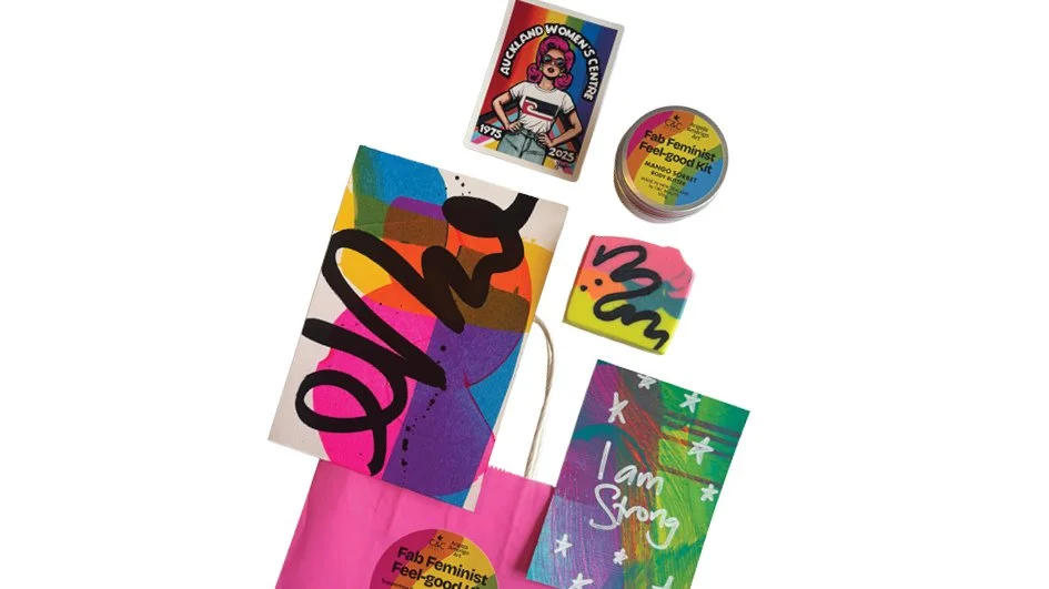 Collection of colorful feminist-themed items, including stickers, a pink bag, and a small jar, with messages like "Queerland Women's Center" and "I am Strong."