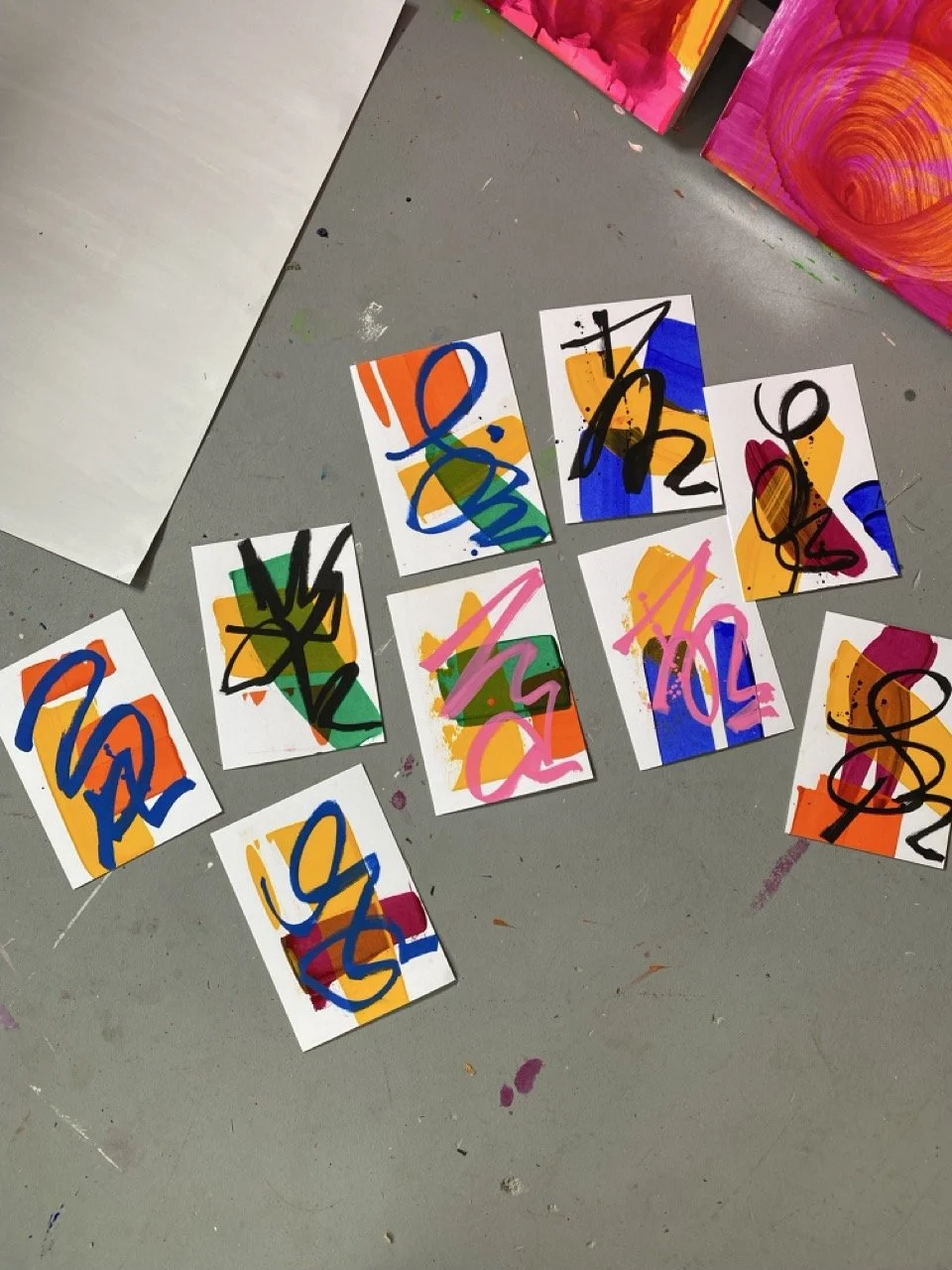 Abstract colorful paintings with bold black scribbles on small white canvases laid out on a gray table, with pink, blue, orange, yellow, and green colors.