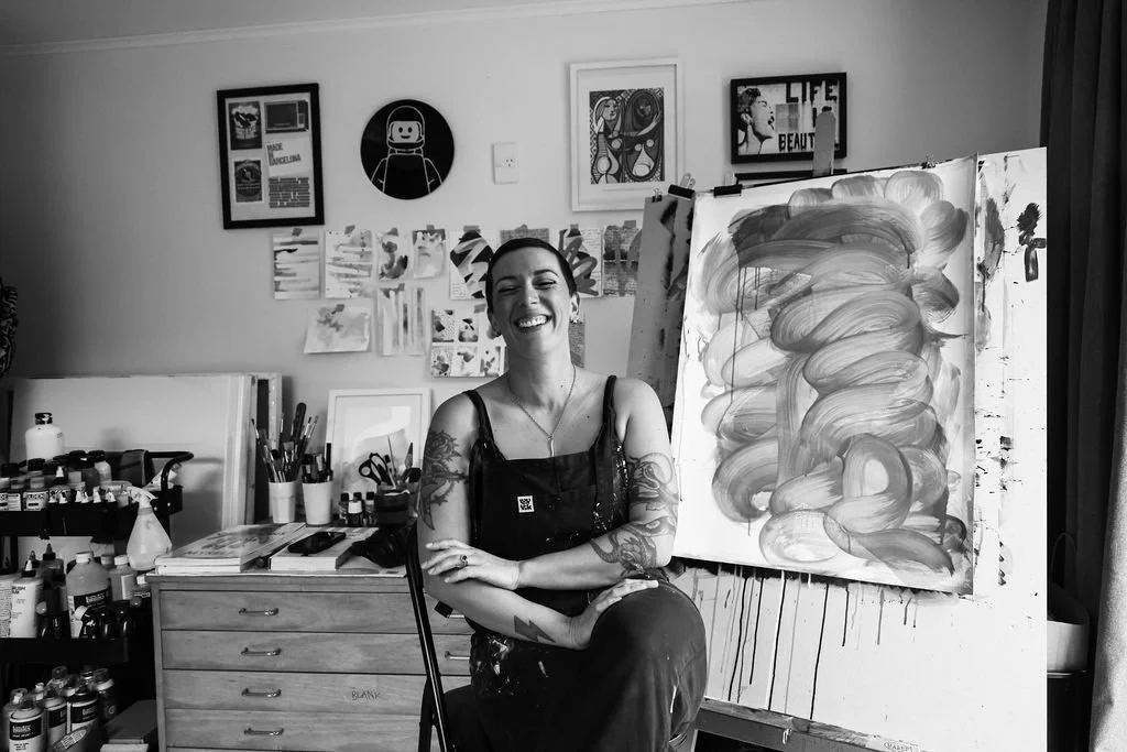 A woman with tattoos smiling and sitting in her art studio next to a large abstract painting on an easel, surrounded by art supplies on a drawer and wall art in the background.