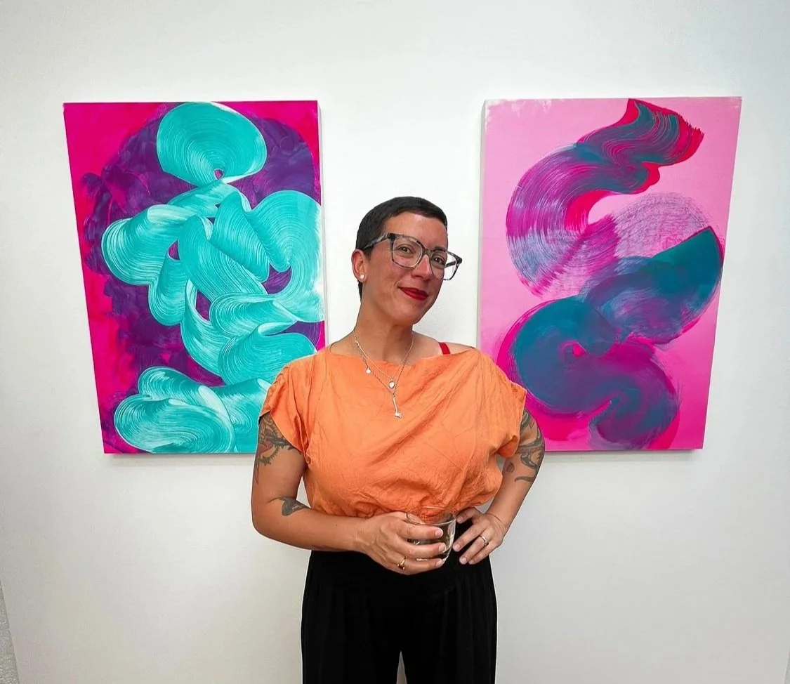 A woman with short dark hair, glasses, and tattoos, wearing an orange top and black pants, standing with a drink in front of two vibrant abstract paintings with pink, blue, purple, and teal brushstrokes.