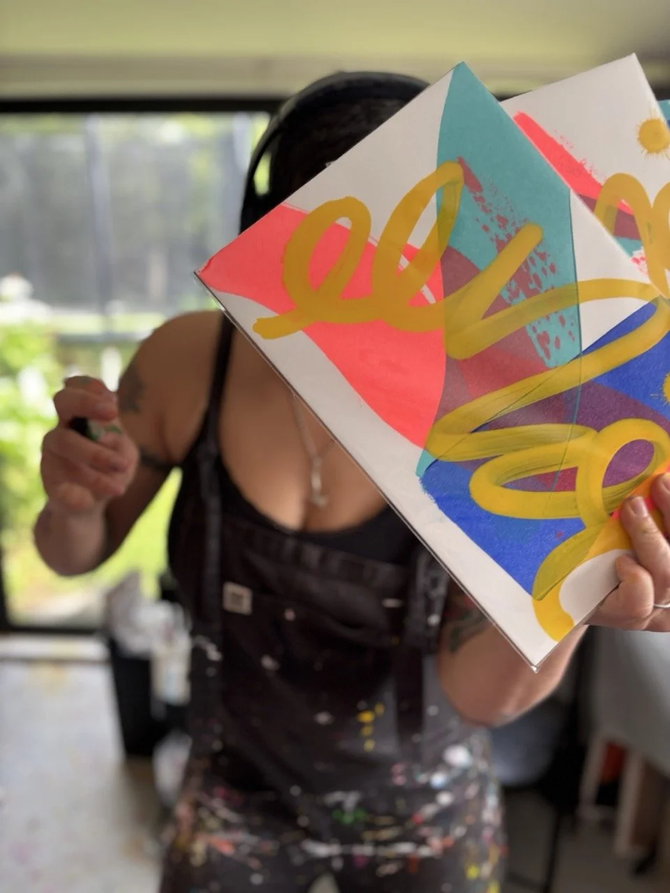 A person holding a colorful, abstract painting or artwork that covers part of their face and torso, with a background of a window and greenery outside.