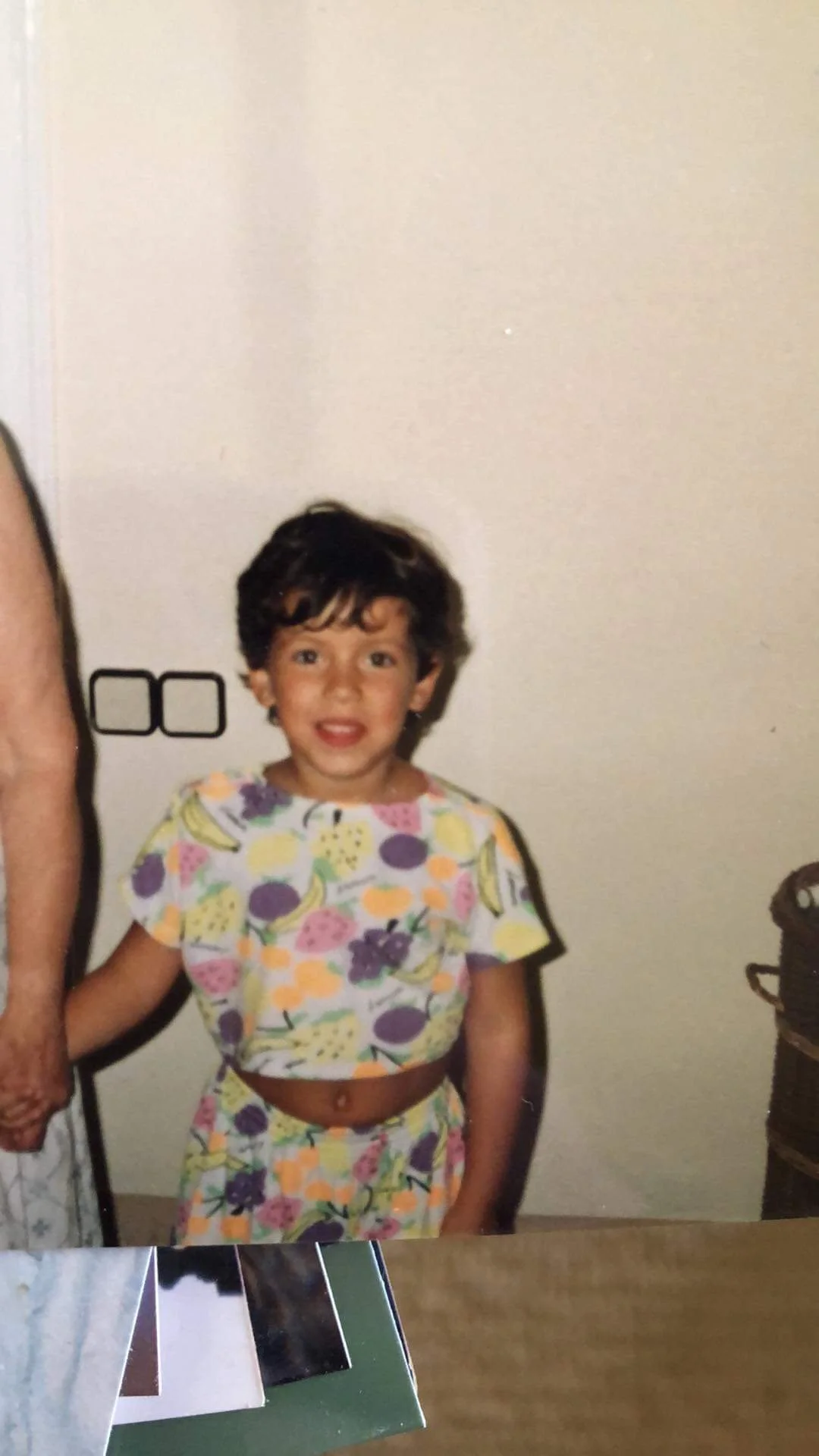 A young girl with curly dark hair, wearing a colorful fruit-themed two-piece outfit, standing indoors against a plain wall. Part of an adult's arm and hand are visible on the left side of the image.