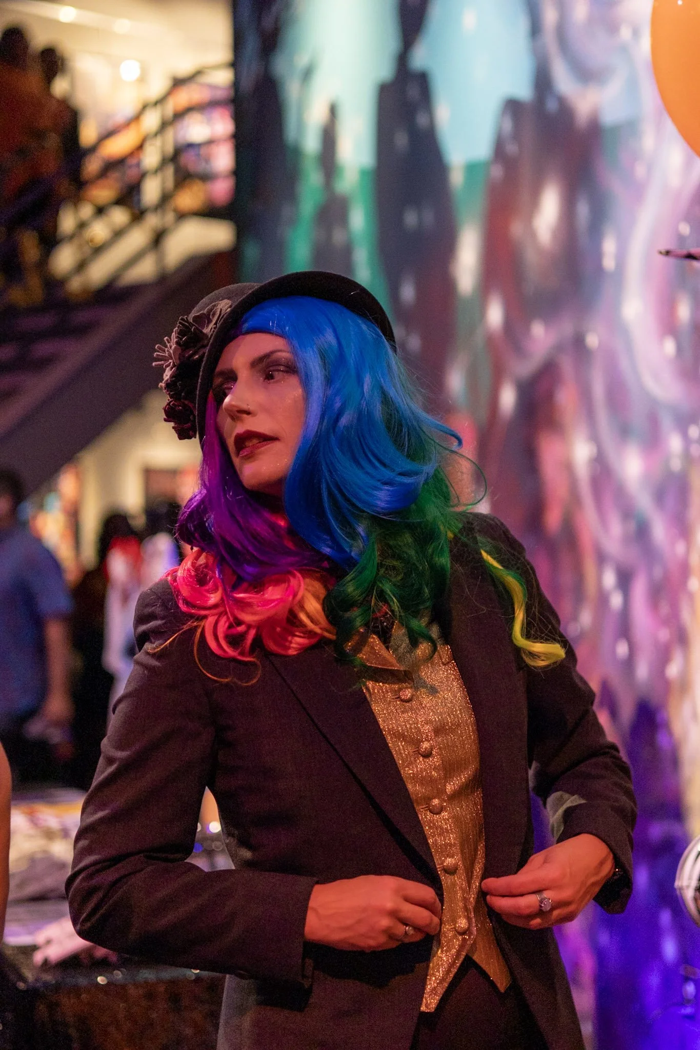 Woman with vibrant rainbow-colored hair, wearing a black hat with a flower, a dark blazer, and a gold top, standing at an indoor event with colorful abstract art in the background.