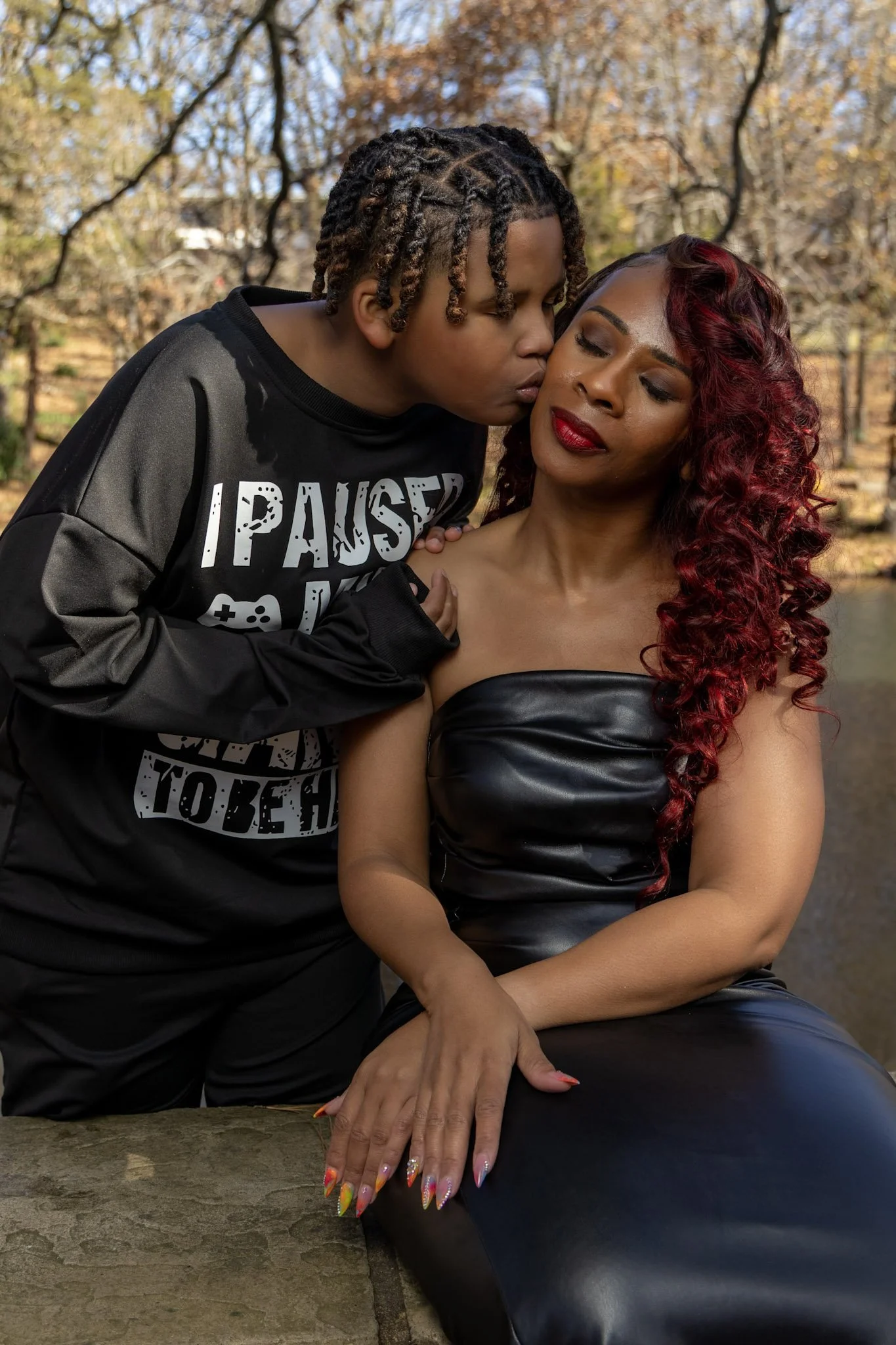 A young person with braided hair kisses a woman with long red curly hair on the forehead while she sits on a rock by a pond in a park with autumn trees.