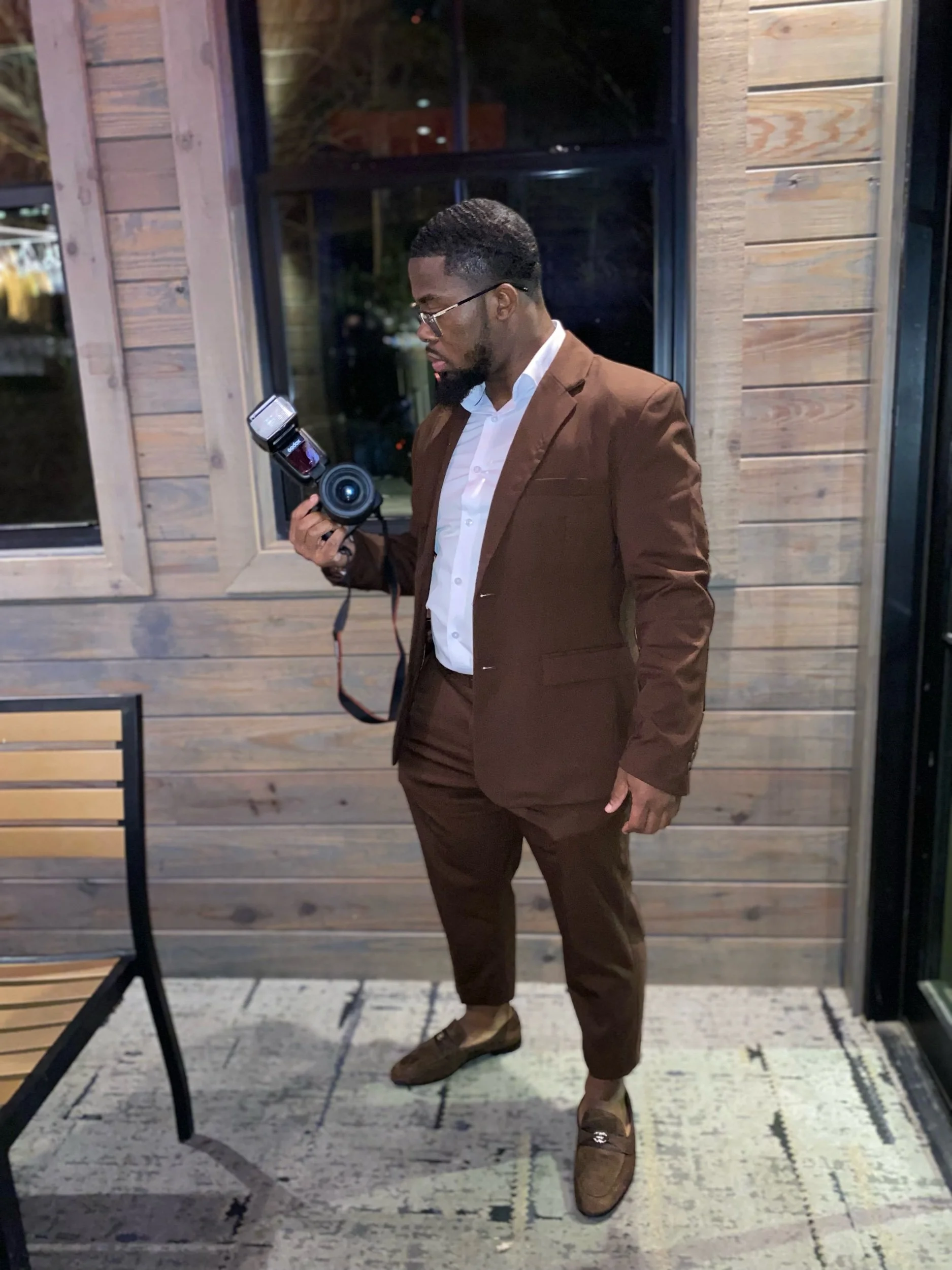 A man in a brown suit and white shirt looking at a camera in his hand, standing outside a wooden building at night.
