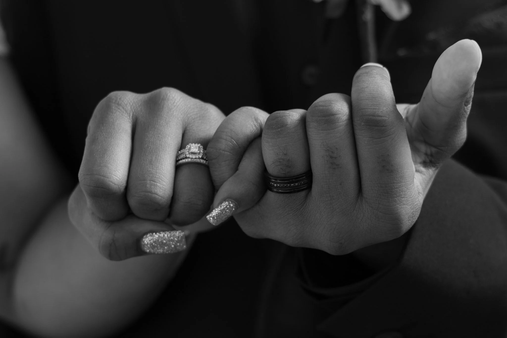 Close-up of two hands clasped together, each wearing wedding rings, with one hand having detailed manicure and the other with darker skin and plain rings, in black and white.