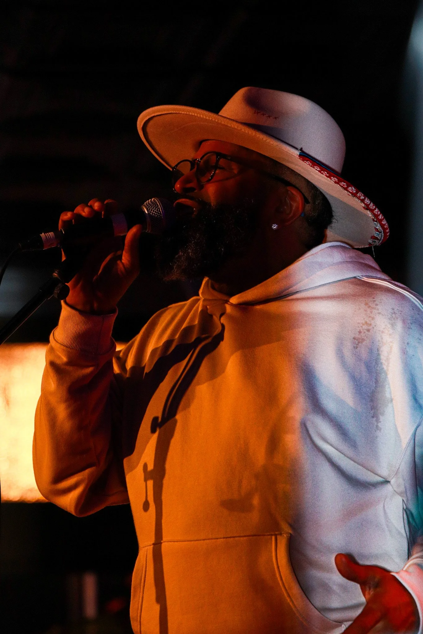 A man with glasses and a beard singing into a microphone, wearing a wide-brimmed hat and a light-colored hoodie, illuminated by stage lighting.