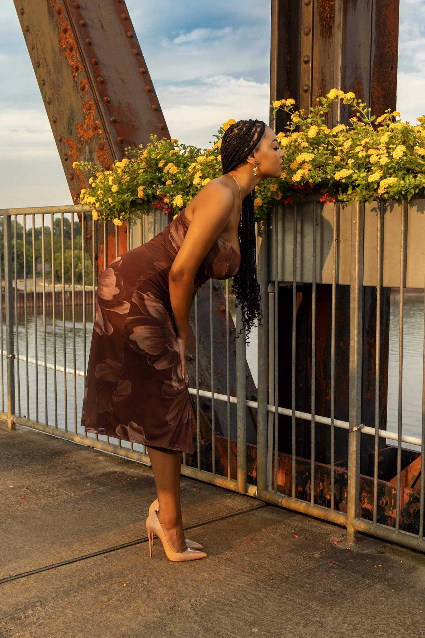 A woman in a brown floral dress and nude high heels leans forward to smell yellow flowers in a rectangular planter on a riverside bridge.