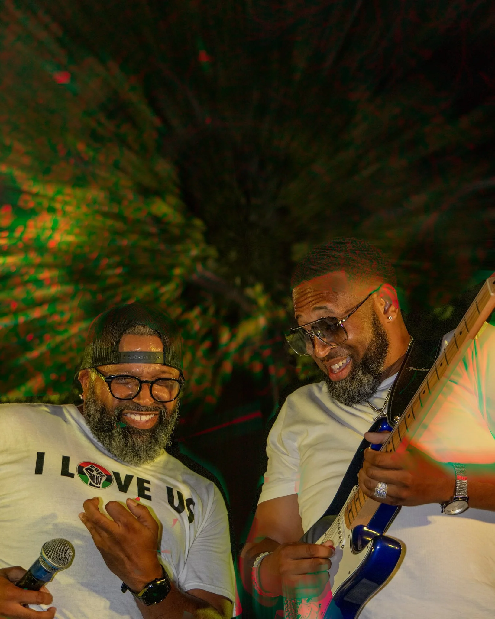 Two men with beards, sunglasses, and white t-shirts smiling and performing with a microphone and an electric guitar outdoors at night with colorful lights in the background.