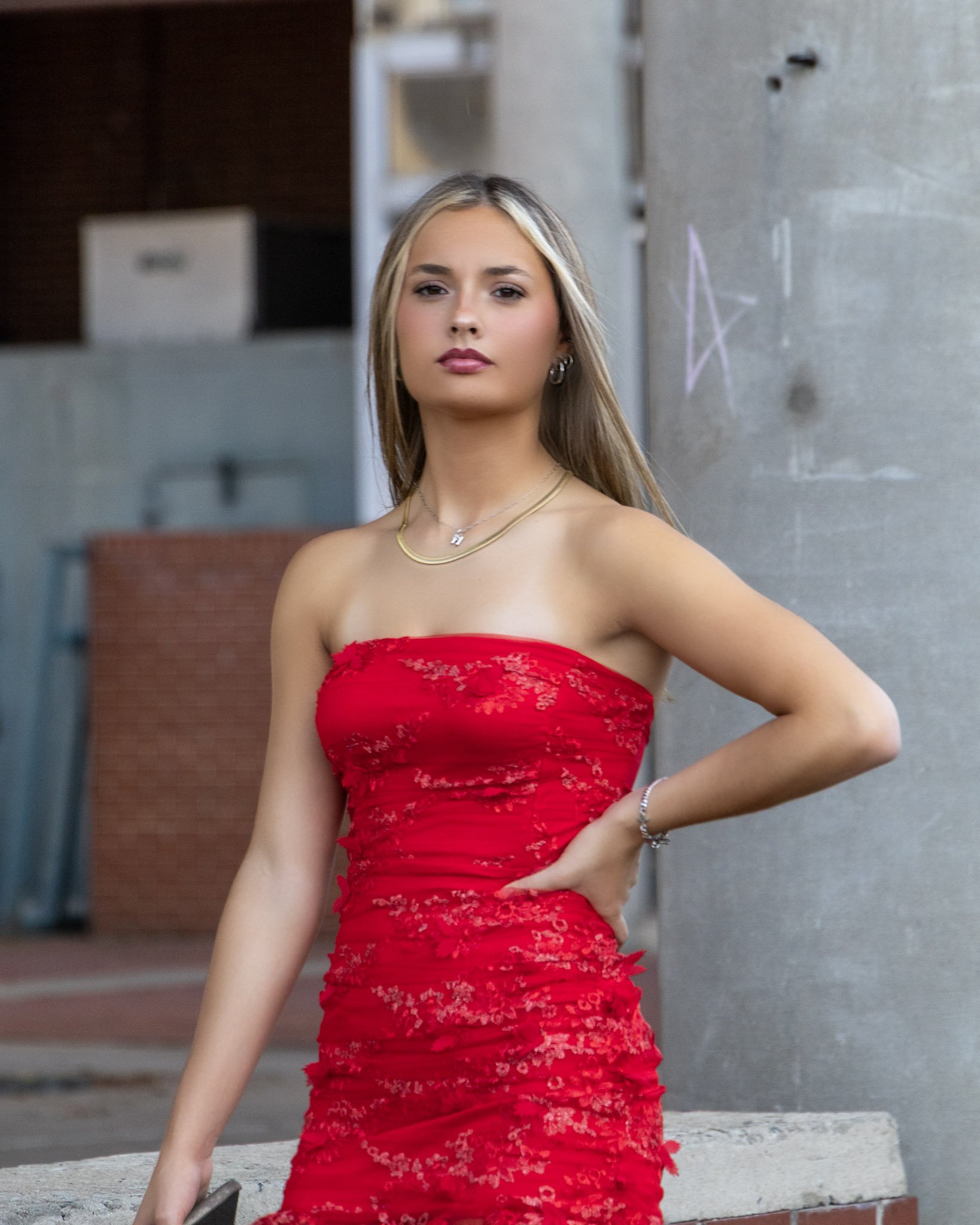 Young woman in a red strapless dress standing outdoors near a concrete wall.