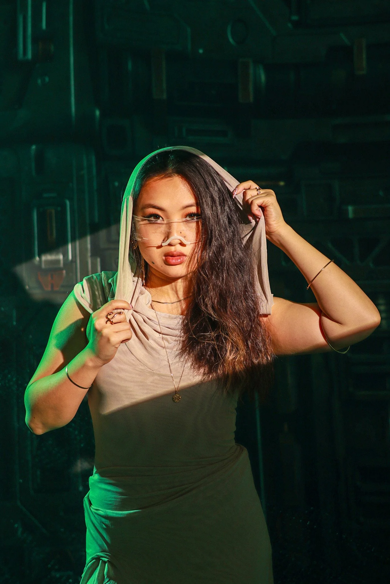 A woman with long curly hair wearing a beige sleeveless top and a necklace, holding a hood over her head, standing in a dark, high-tech setting with green lighting.