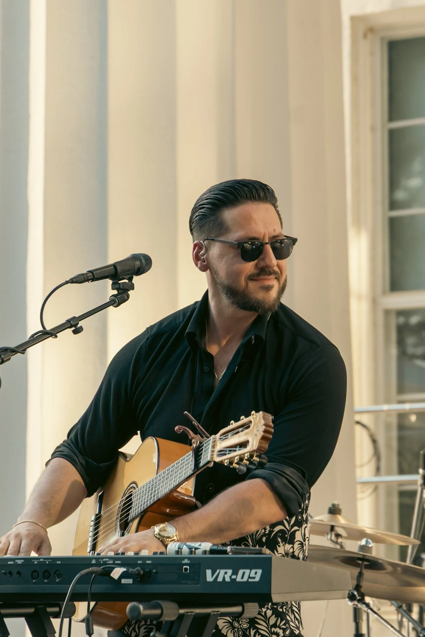 A man wearing sunglasses plays an acoustic guitar and a keyboard during a performance.