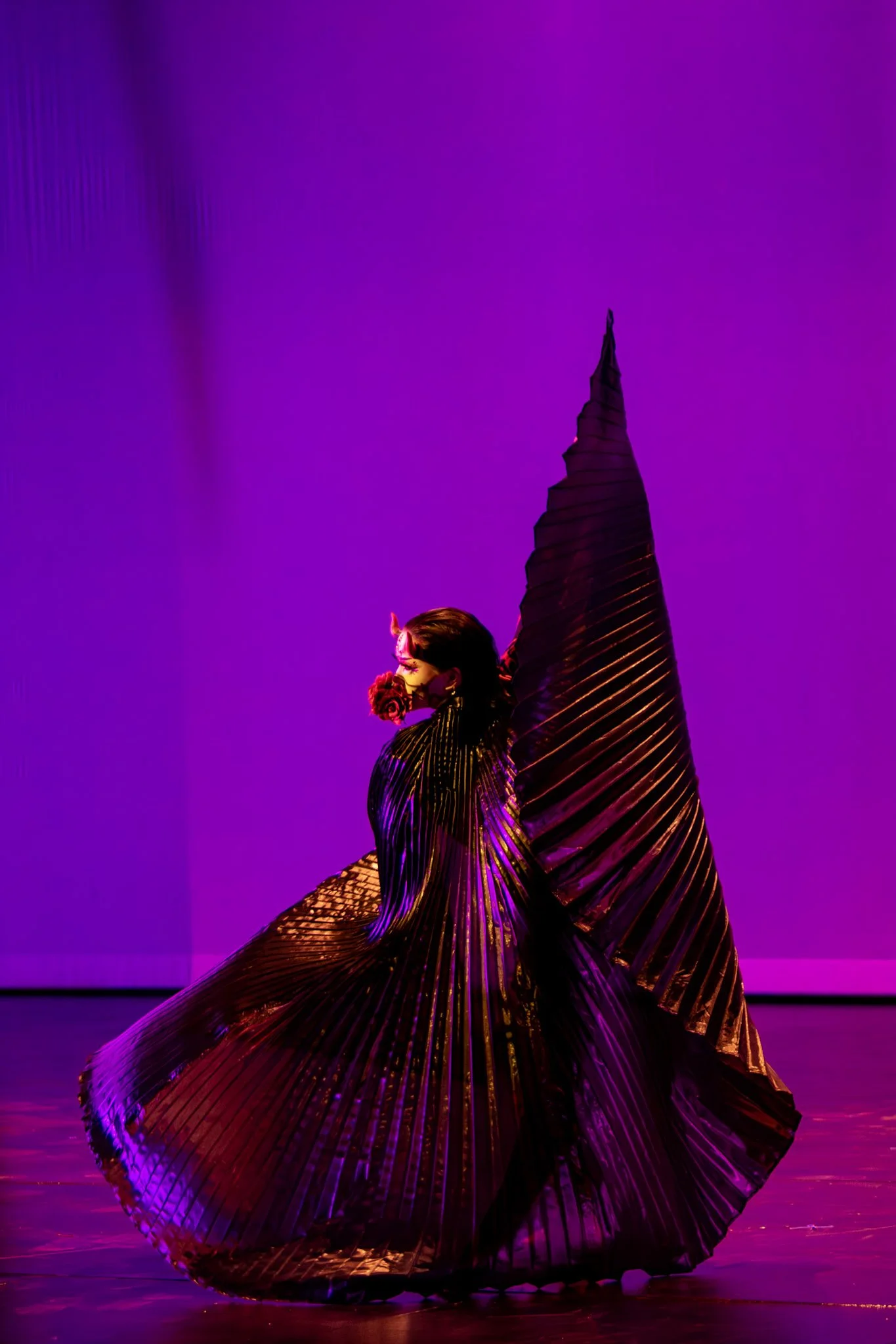 A performer in an elaborate black and purple costume with large, pleated fabric accents, wearing a mask and headpiece, performing on a stage with purple lighting.