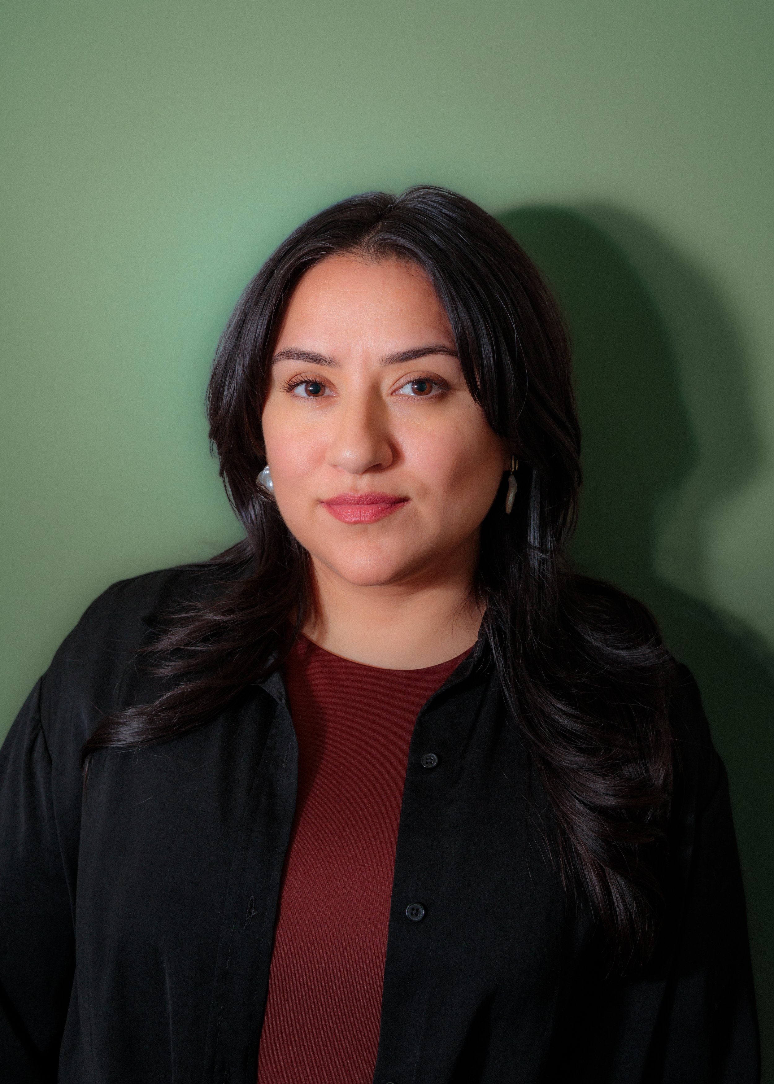 Portrait of a professional woman of color with dark hair, wearing a maroon shirt and black jacket, standing against a green background.