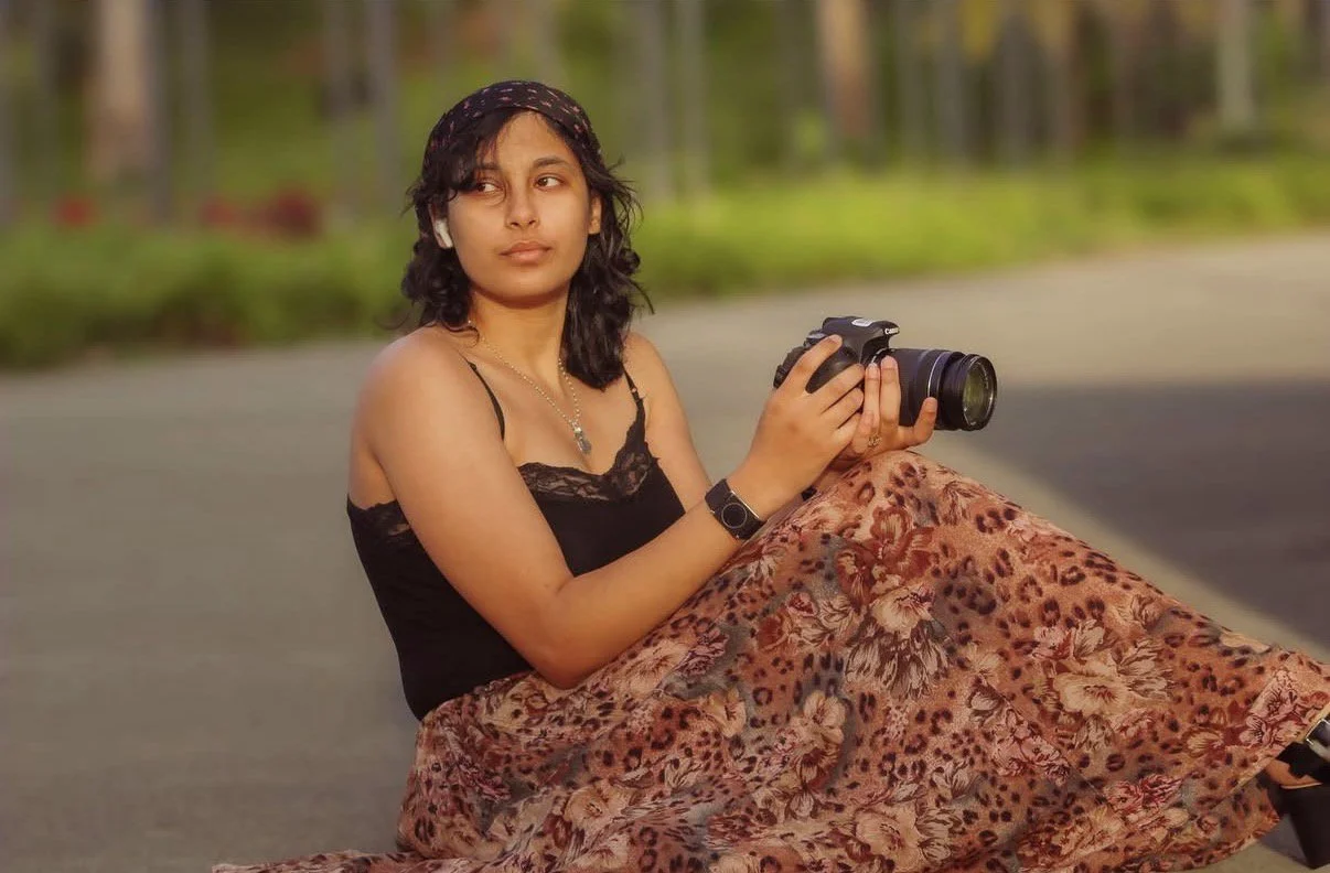 A photo of me sitting on the ground outdoors, holding a camera, with trees and a blurred background.