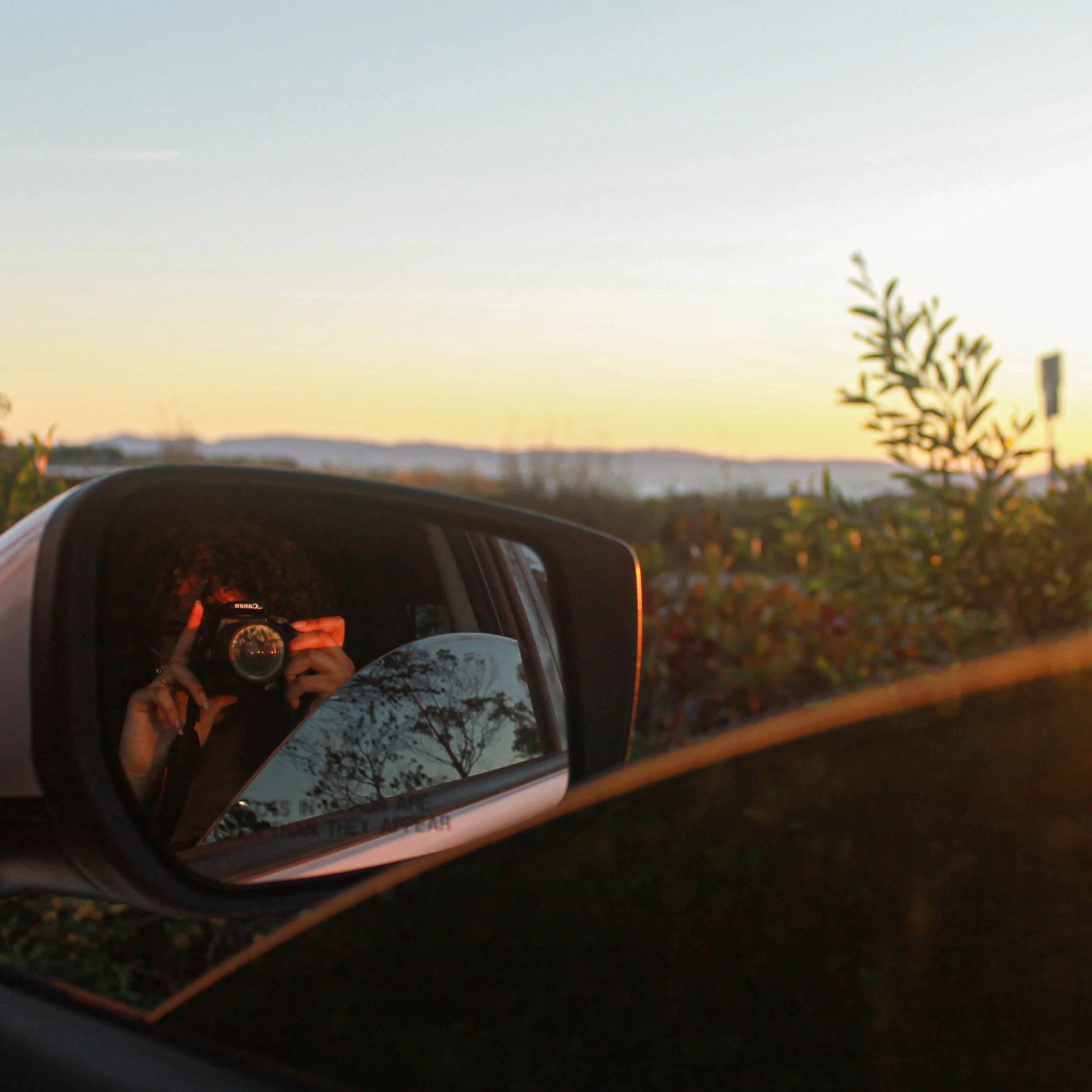 View of a sunset with a side mirror of a car reflecting myself taking a photo with a camera, outdoors with trees and bushes in the background.