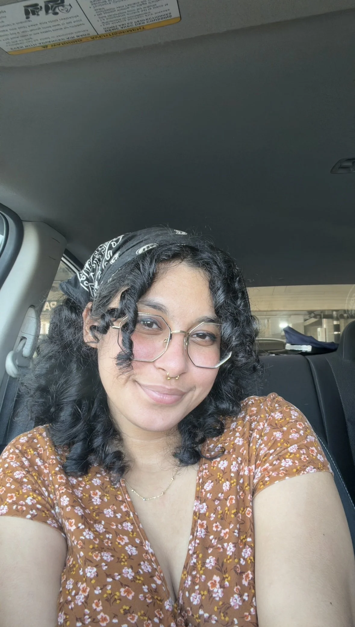 A photo of me with glasses and a bandana, smiling inside a car, wearing a floral brown top and a delicate necklace.